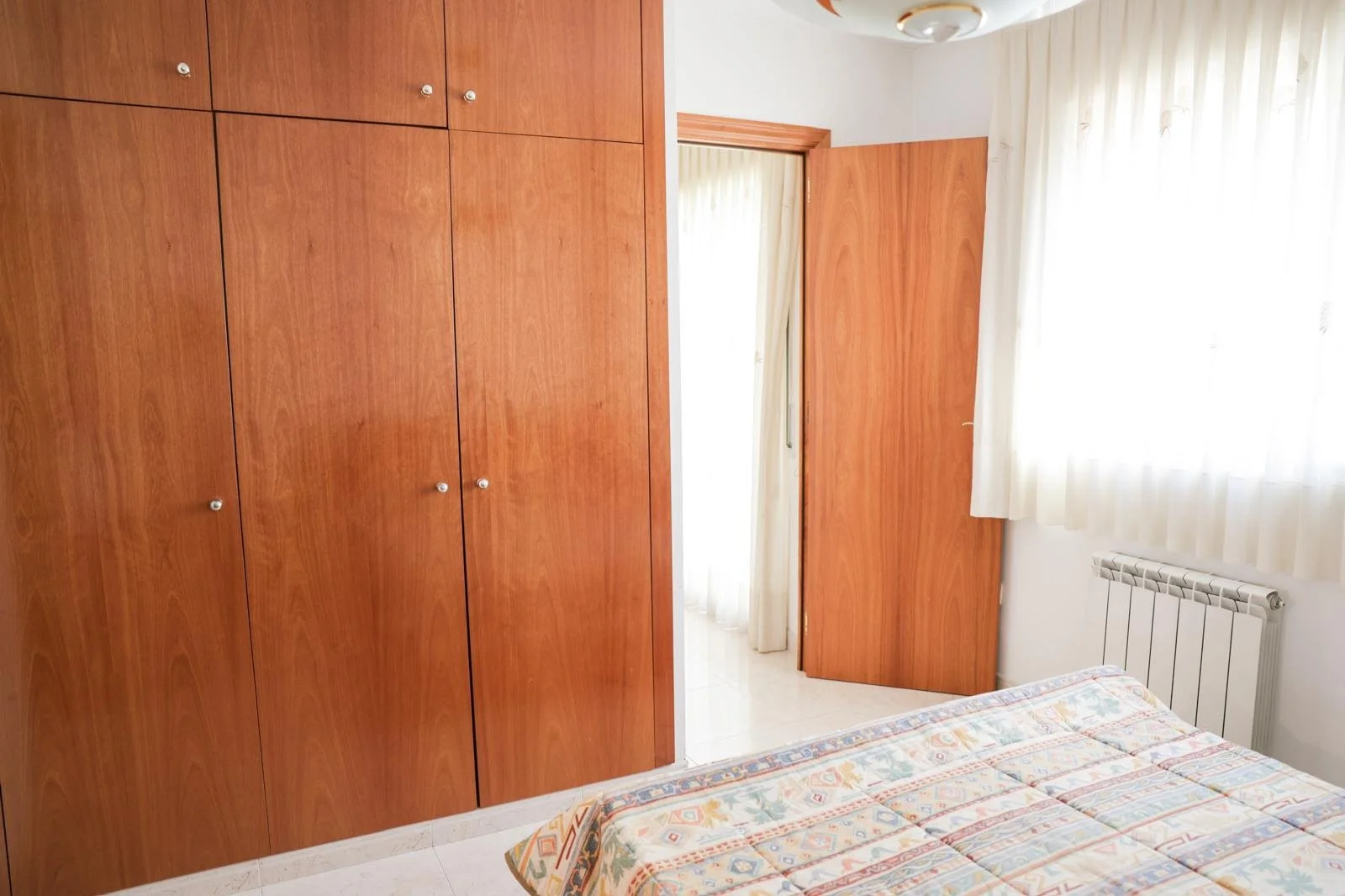 Habitación con armario de madera y cama con colcha colorida, ventana con cortinas blancas y radiador.
