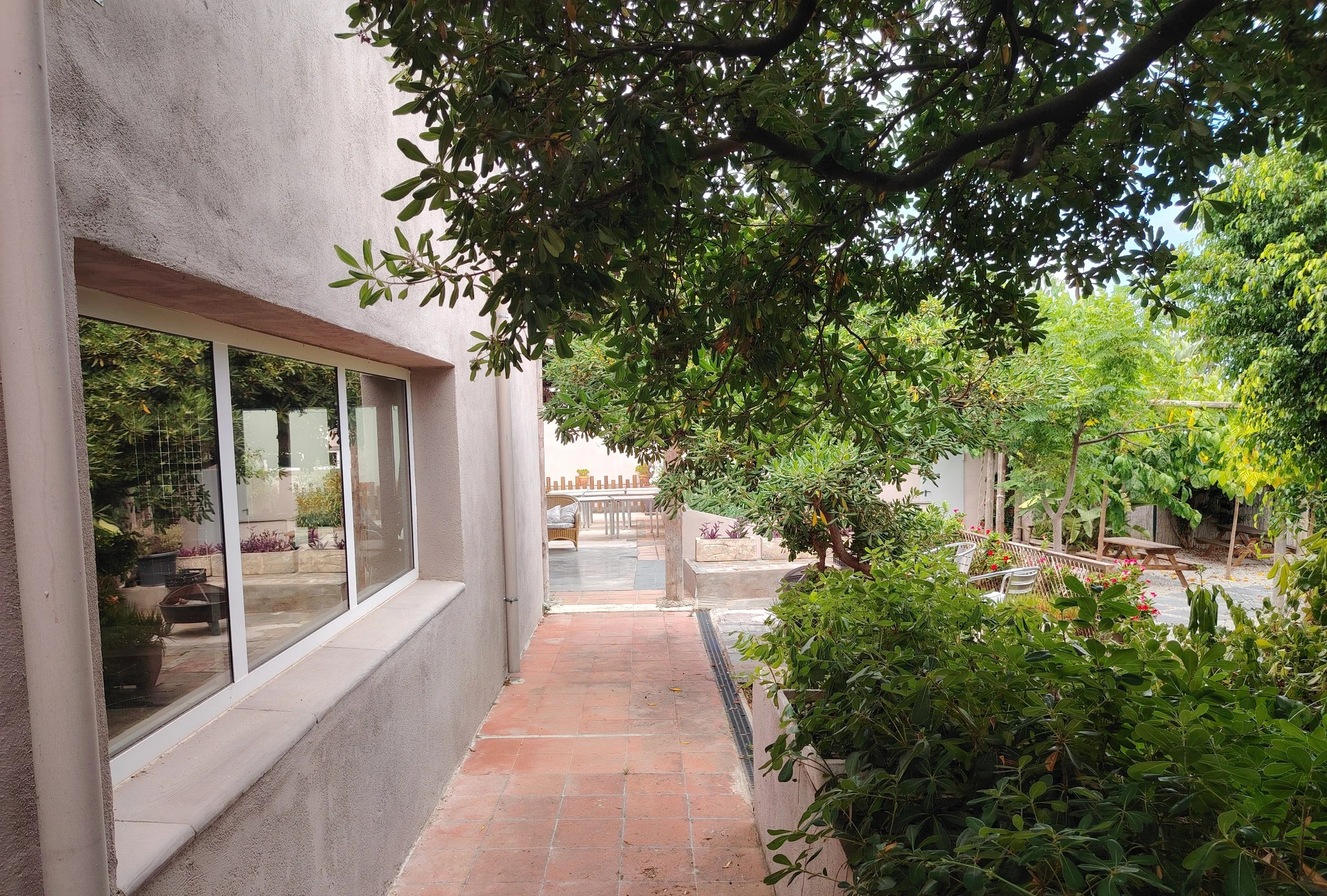 Jardín con árboles y plantas, vista de un espacio exterior con mobiliario y cercas, al lado de una casa con ventanas.