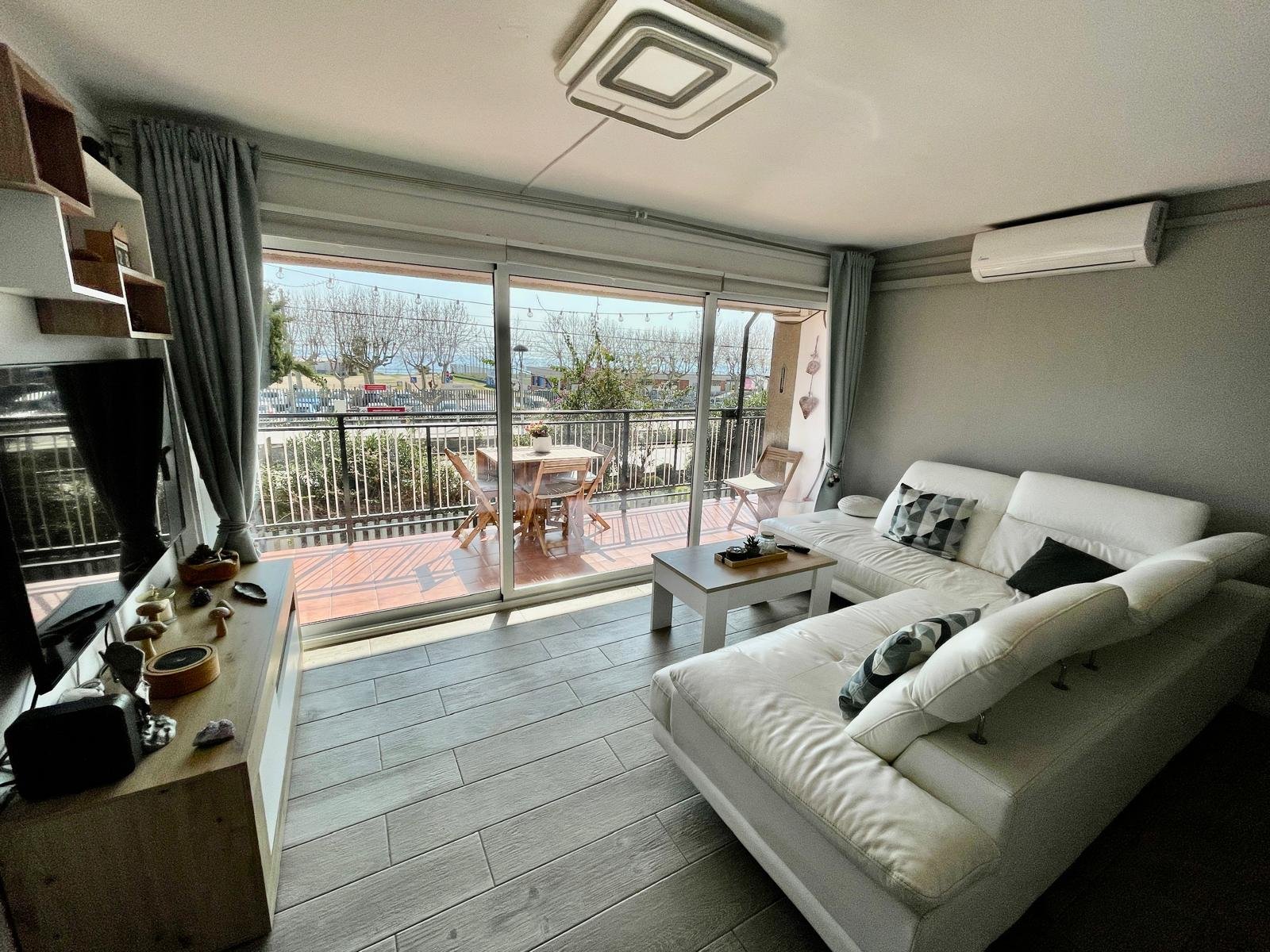 Sala de estar con sofá blanco, mesa de centro, televisión, y un balcón con terraza y muebles exteriores, con vista a árboles y una calle.