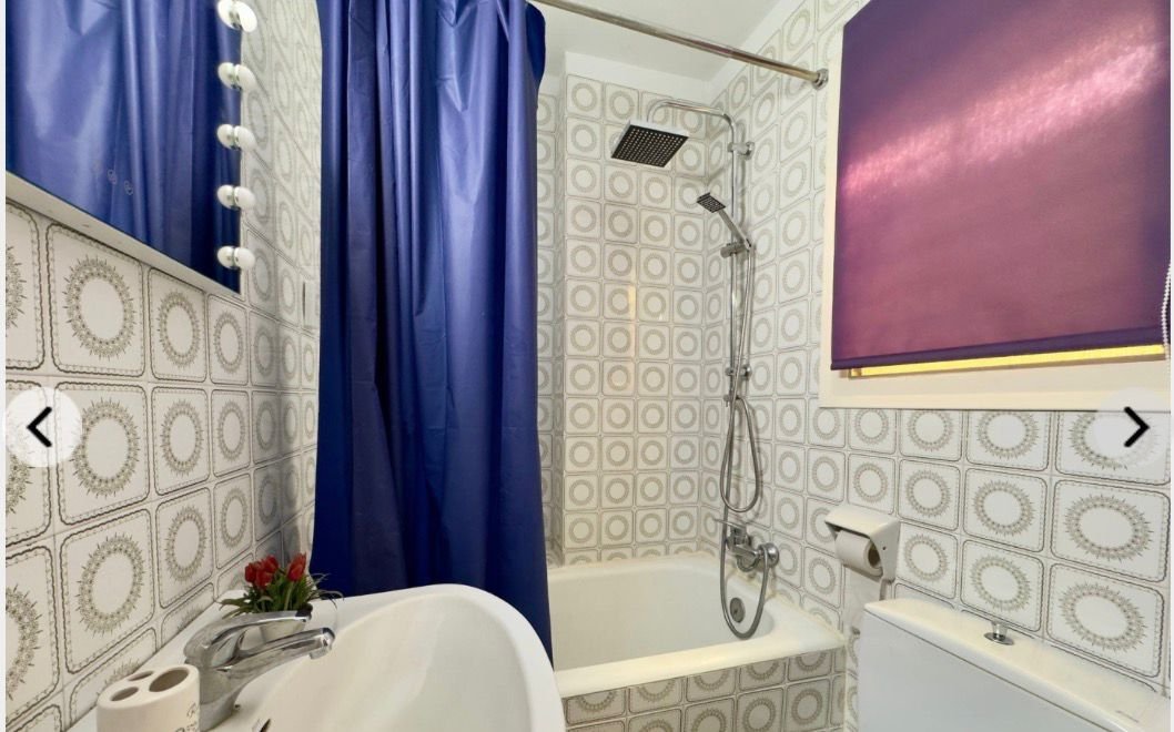 Baño con azulejos de diseño geométrico, cortina azul, ducha con cabezal de lluvia, ventana con persiana morada, lavabo con flor artificial, papel higiénico, inodoro.