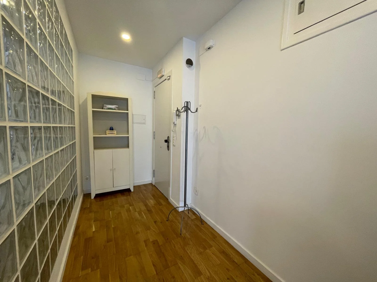 Vestíbulo con parquet de madera, estantería blanca, lámpara de pie, pared blanca, puerta de entrada, y muros decorados con bloques de vidrio.