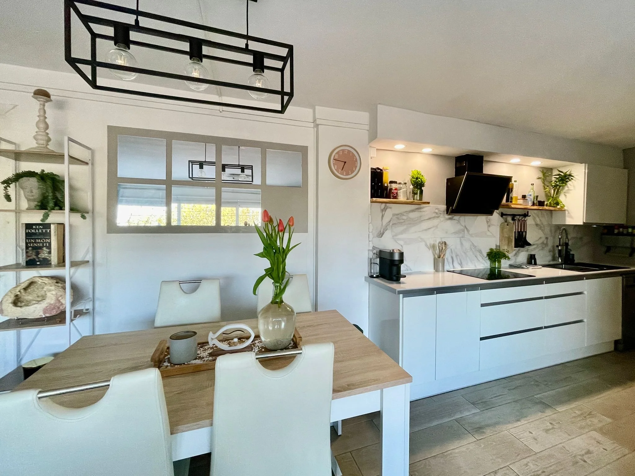 Interior de una sala de comedor con una mesa de madera, sillas blancas, un jarrón con tulipanes en el centro, y fondo que muestra una cocina moderna con gabinete blanco, en la pared un reloj, estantería con libros y plantas.
