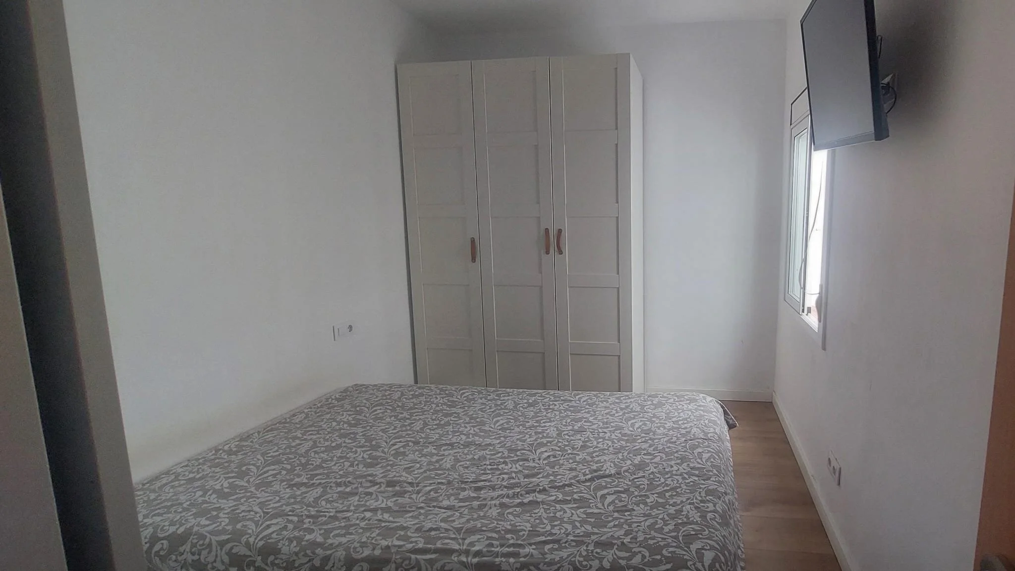 Cuarto con una cama de diseño floral en primer plano, un armario de color gris claro en la pared, una ventana con cortina blanca y una televisión en la esquina derecha.