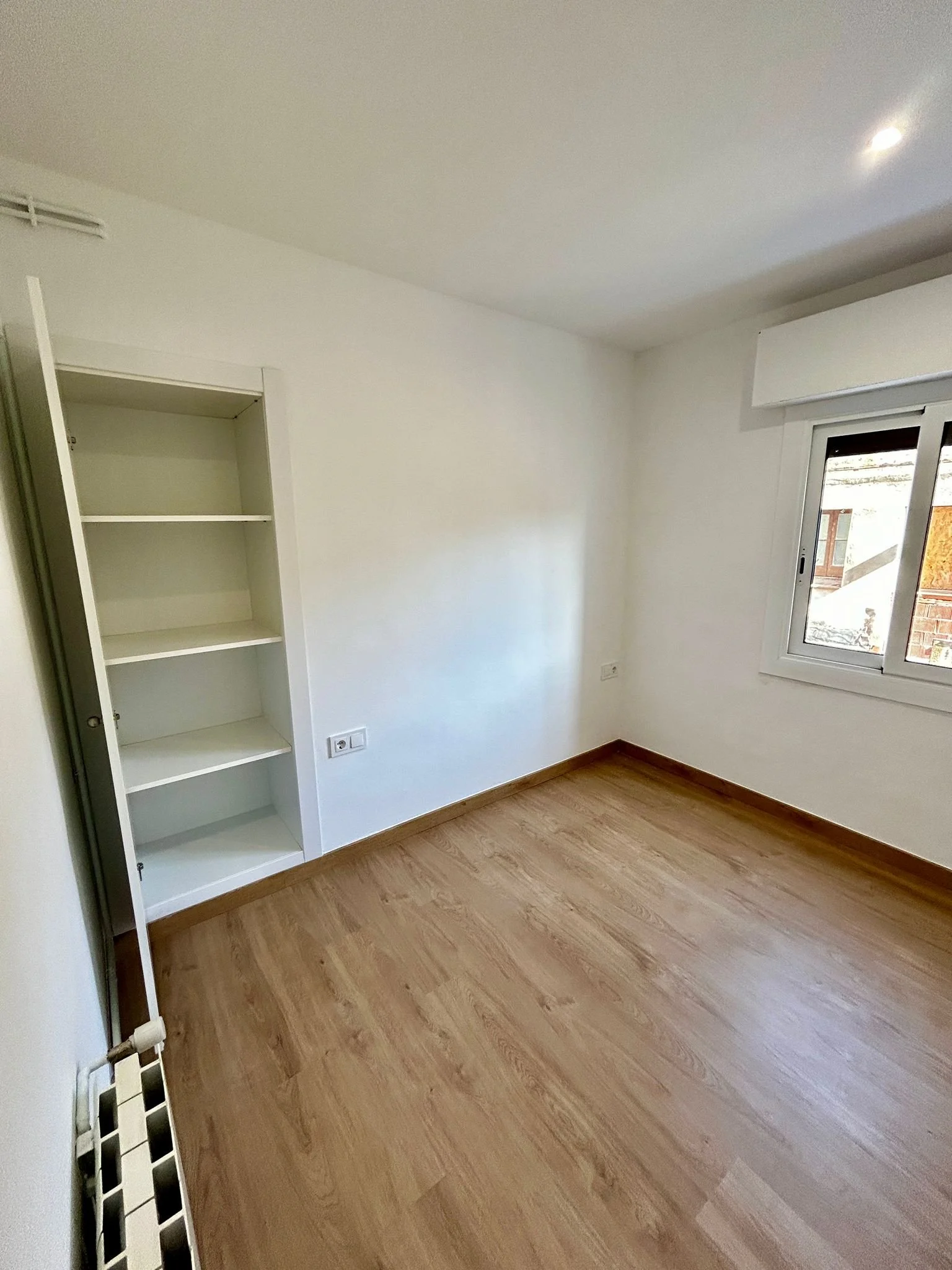 Habitación vacía con paredes blancas, piso de madera, una ventana y una estantería blanca.