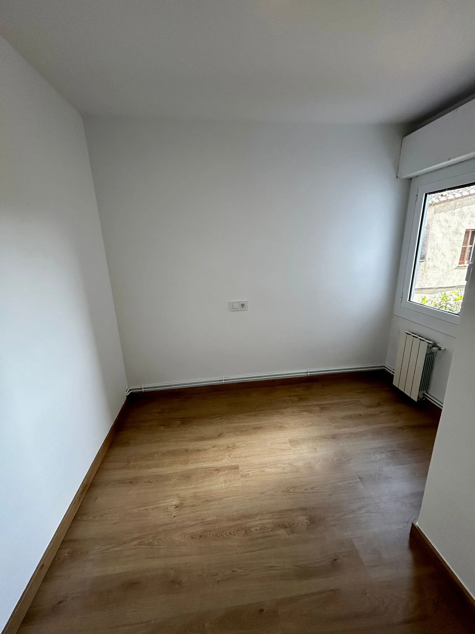 Habitación vacía con paredes blancas, suelo de madera y una ventana grande que permite la entrada de luz natural.