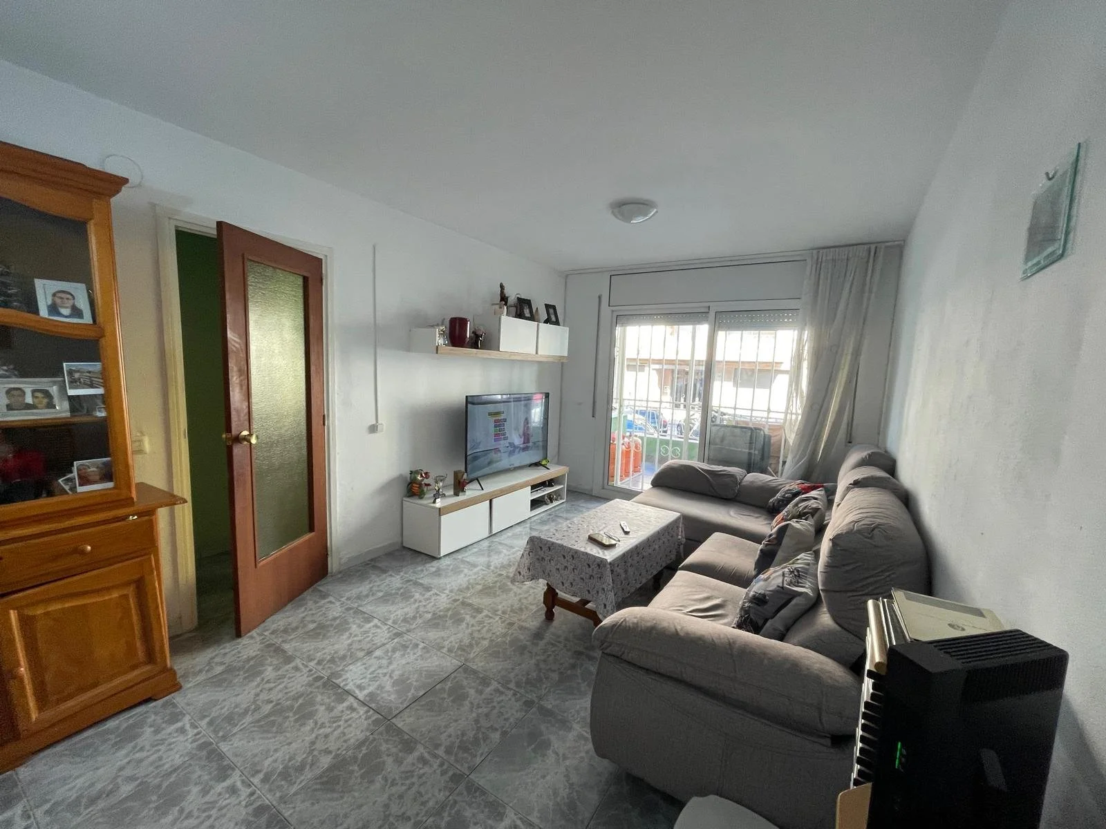 Sala de estar con sillón gris, televisión en mueble blanco, mesa con mantel, ventana grande con cortinas y barredora en balcón.