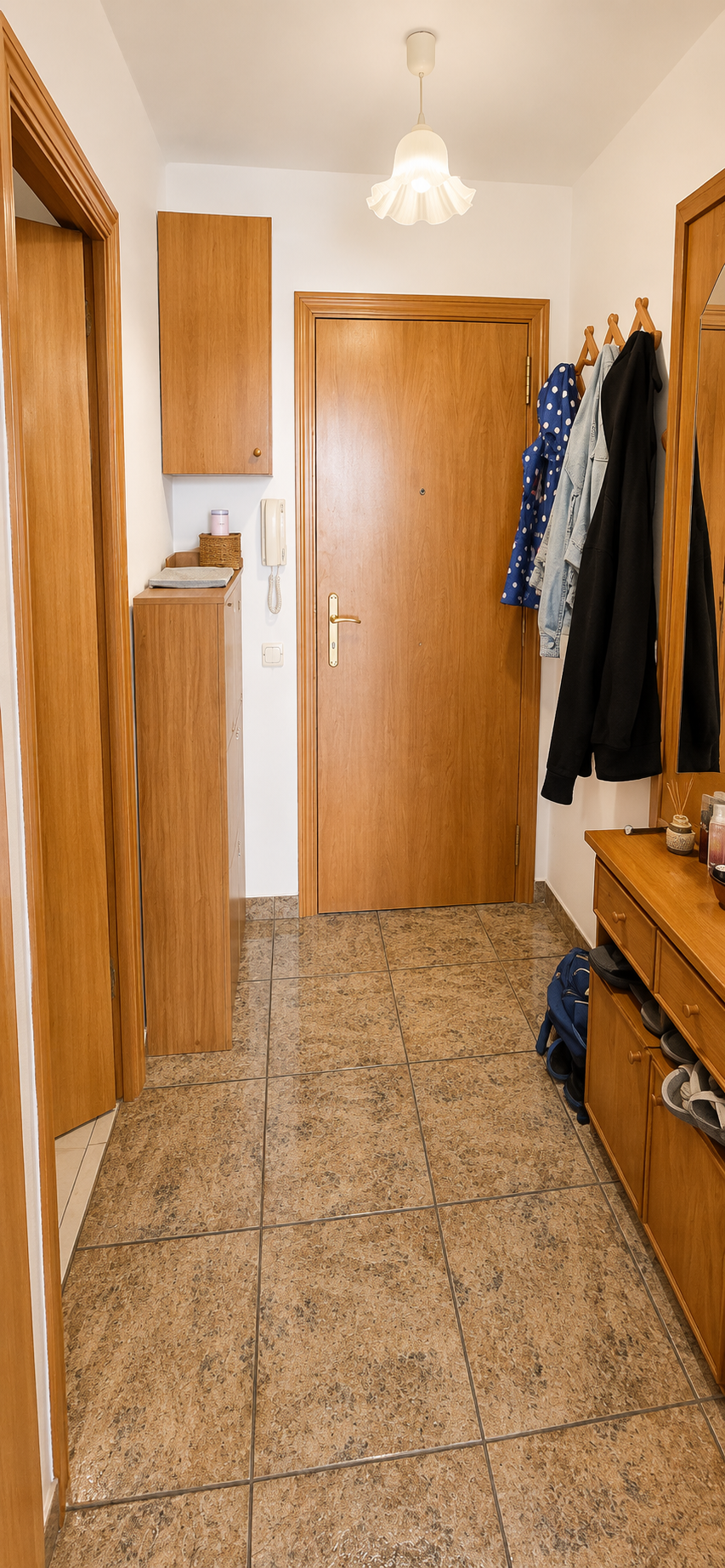 Vestíbulo con piso de baldosas, puertas de madera, armario y perchero con ropa, espejo a la derecha y lámpara en el techo.