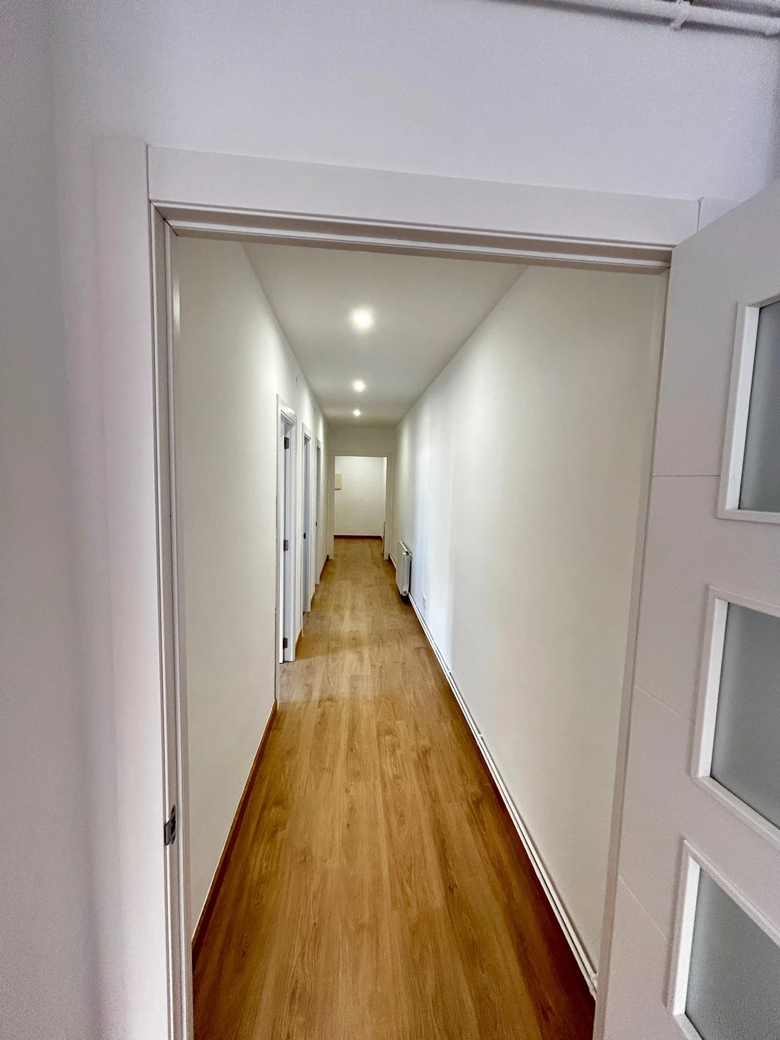 Pasillo largo y estrecho con puertas blancas a cada lado, paredes blancas y piso de madera, con iluminación en el techo.