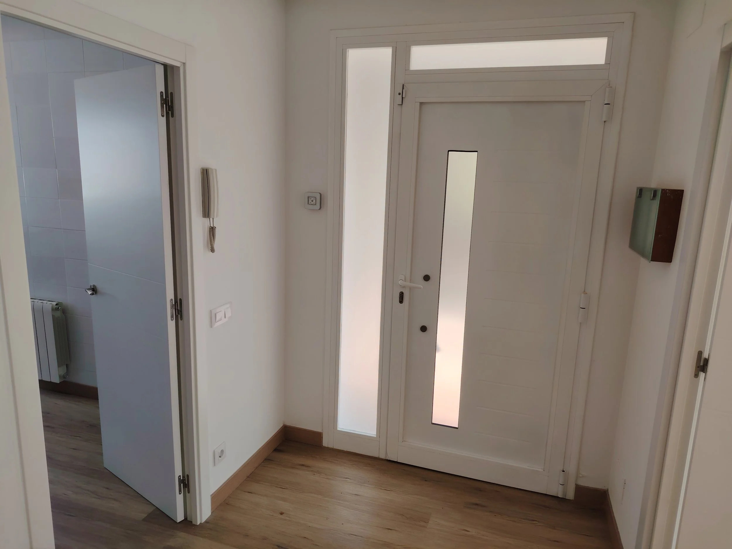 Vestíbulo con puerta de entrada blanca, ventana vertical en la puerta, piso de madera clara, teléfono de pared y caja de correos.