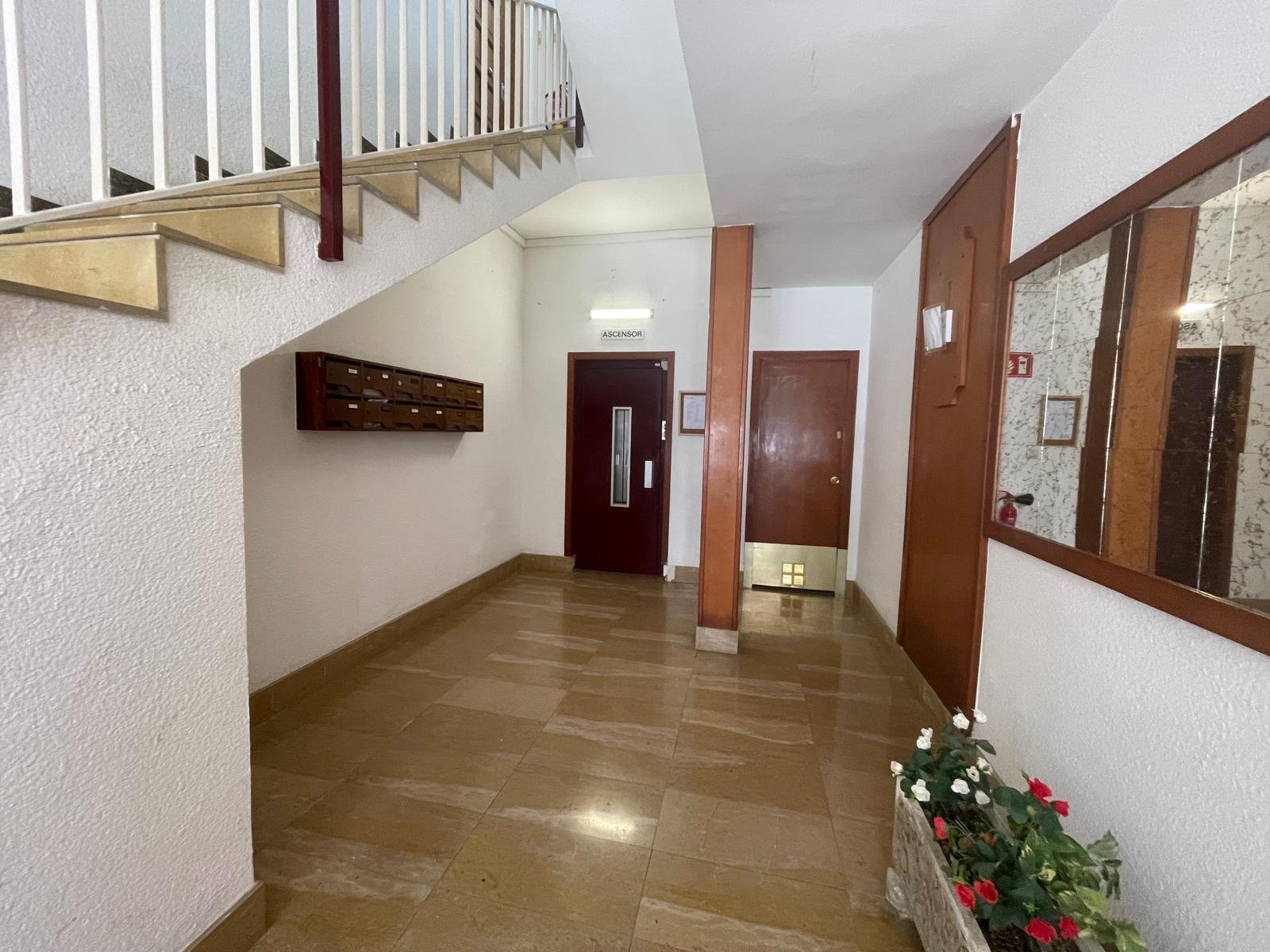 Vestíbulo de un edificio con piso de madera, buzones en la pared izquierda, puerta de ascensor y puerta en la esquina, y un espejo grande en la pared derecha, con flores en una maceta junto a la pared.