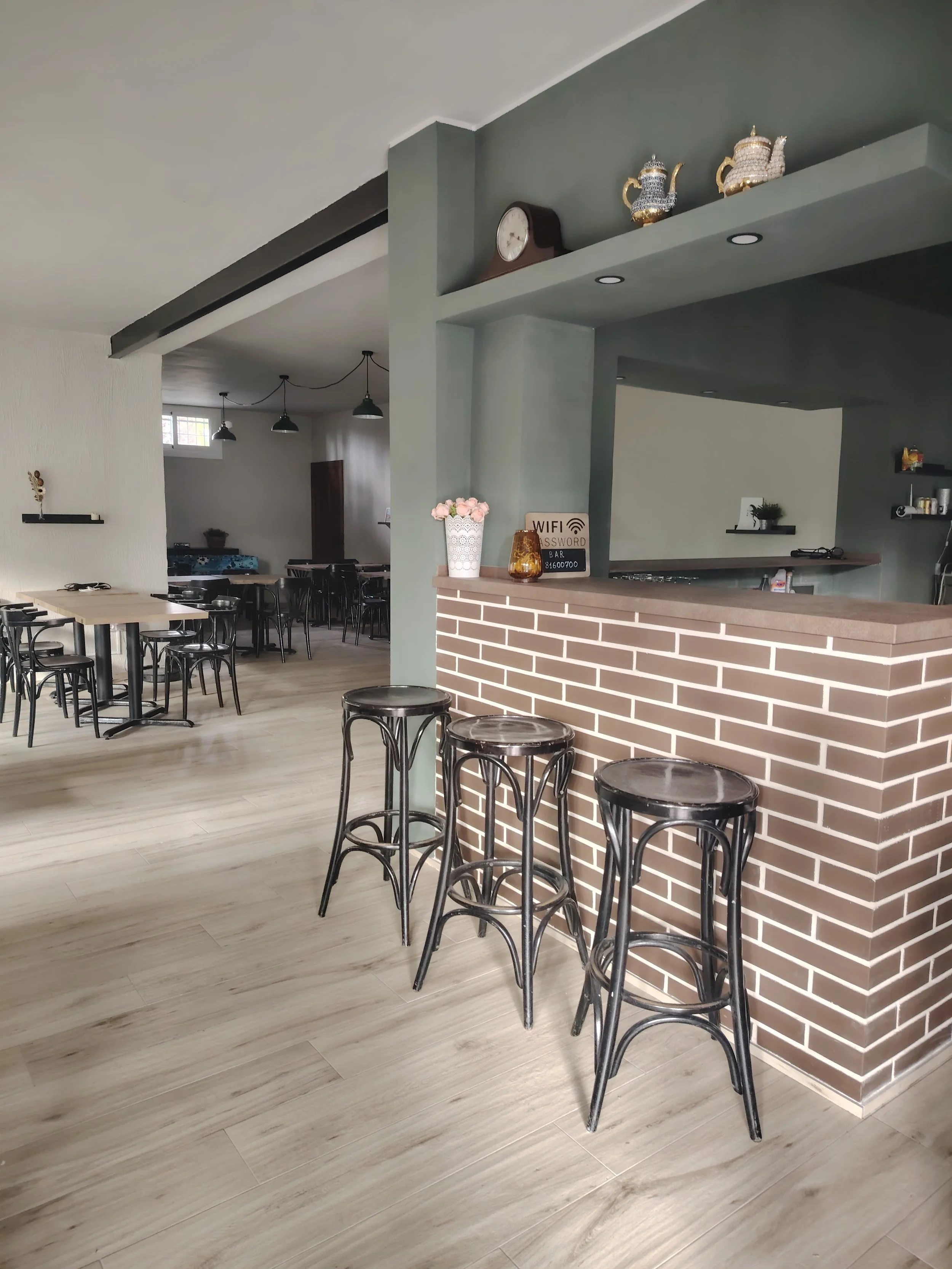 interior de un café con barra de ladrillos, tres sillas altas negras, mesas y sillas negras, decoración minimalista y plantas en las estanterías