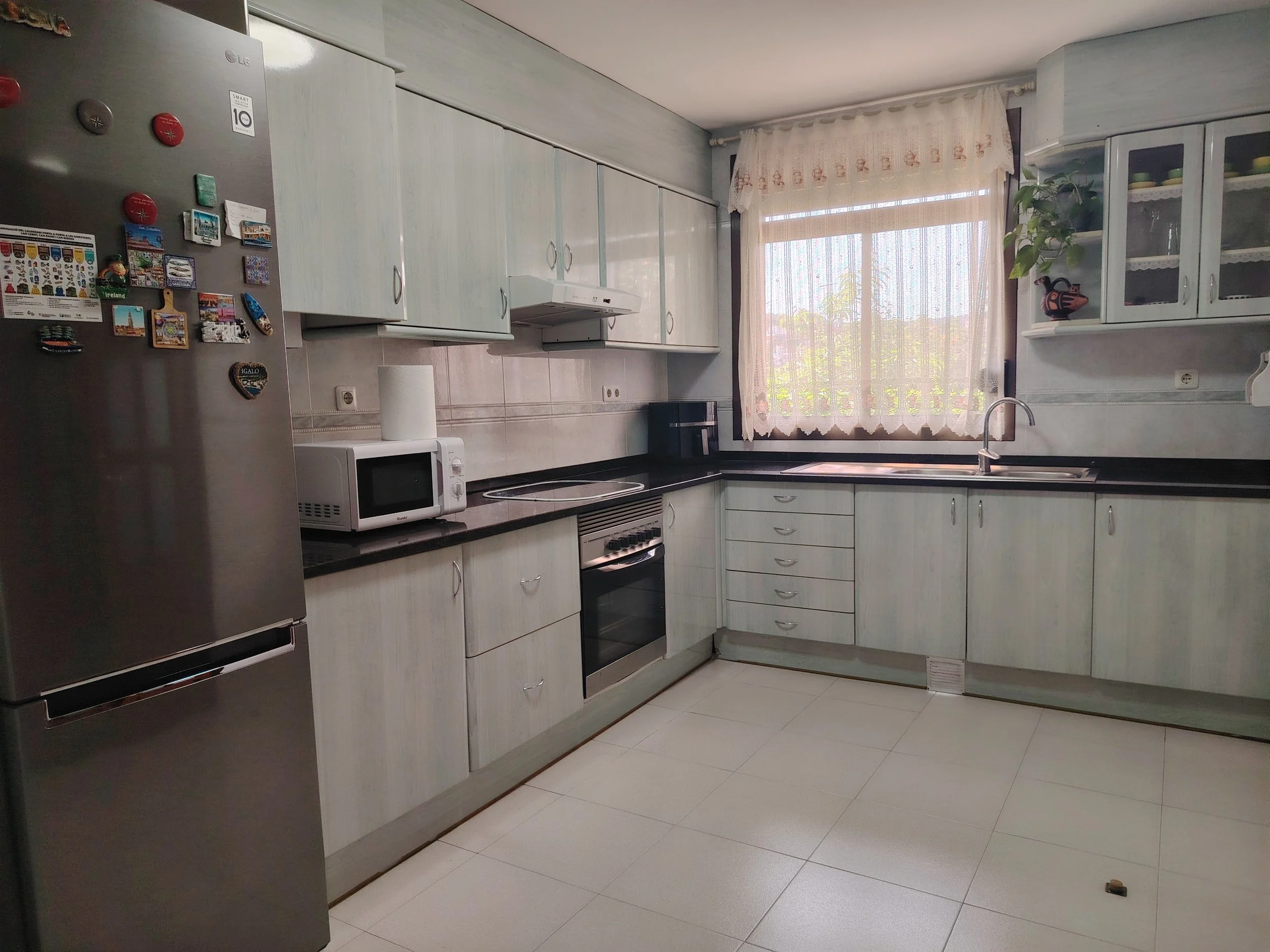Cocina con gabinetes blancos, estufa, microondas, nevera y un fregadero con cortinas blancas en la ventana.
