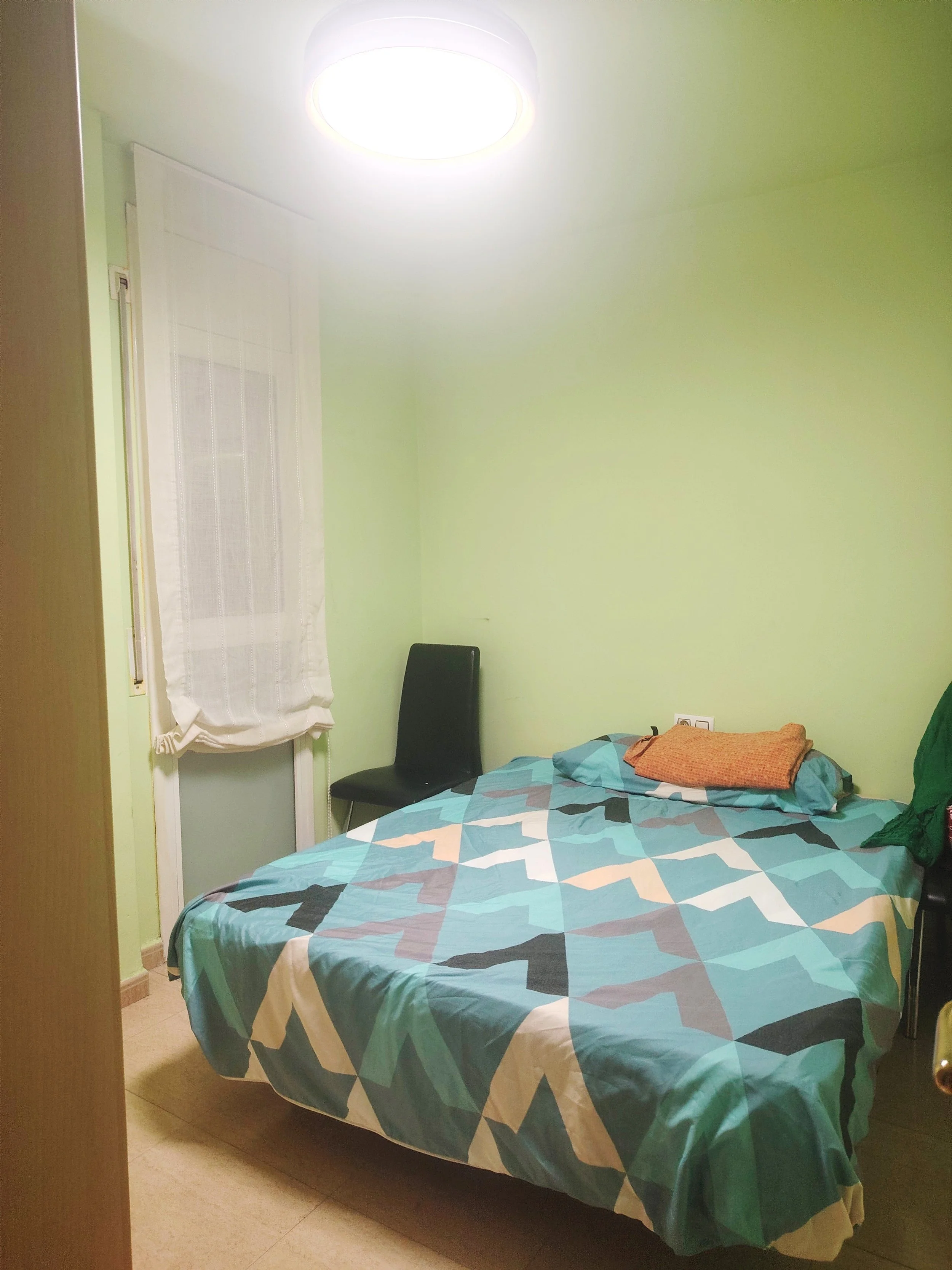 Dormitorio con cama, edredón de colores azules, verde y beige, una almohada naranja, una pared verde, una ventana con cortina blanca, una silla negra junto a la cama, y una lámpara de techo encendida.