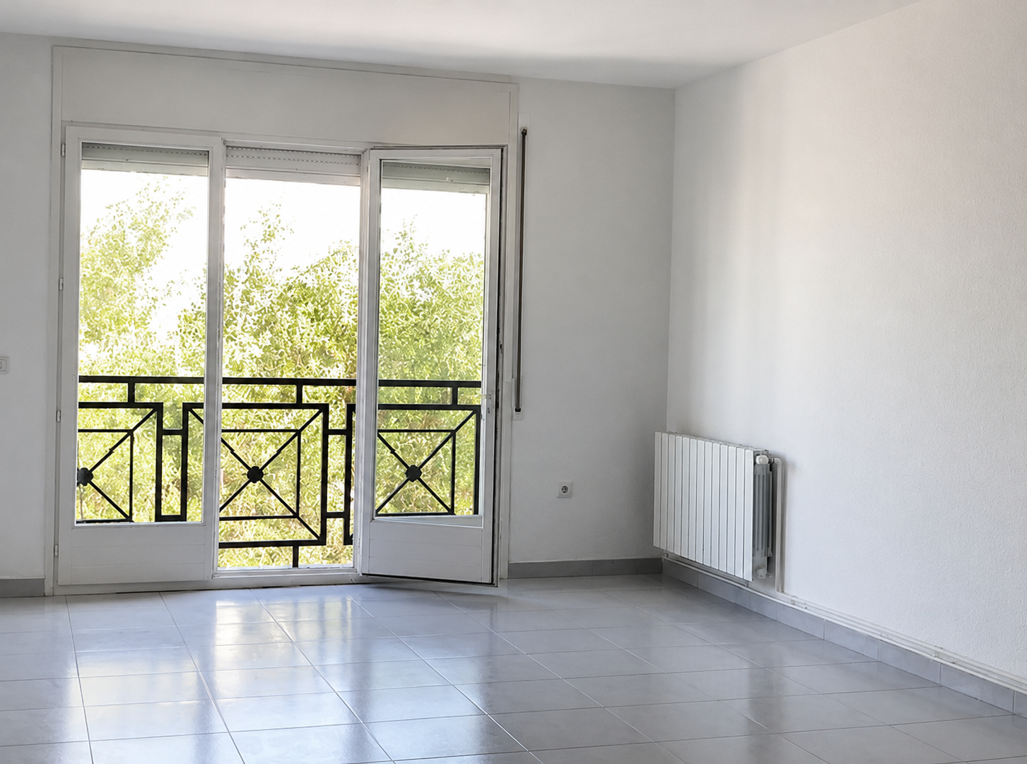 Sala vacía con puerta de balcón de vidrio que da a un balcón con barandilla negra, paredes blancas, radiador blanco y piso de baldosas grises.