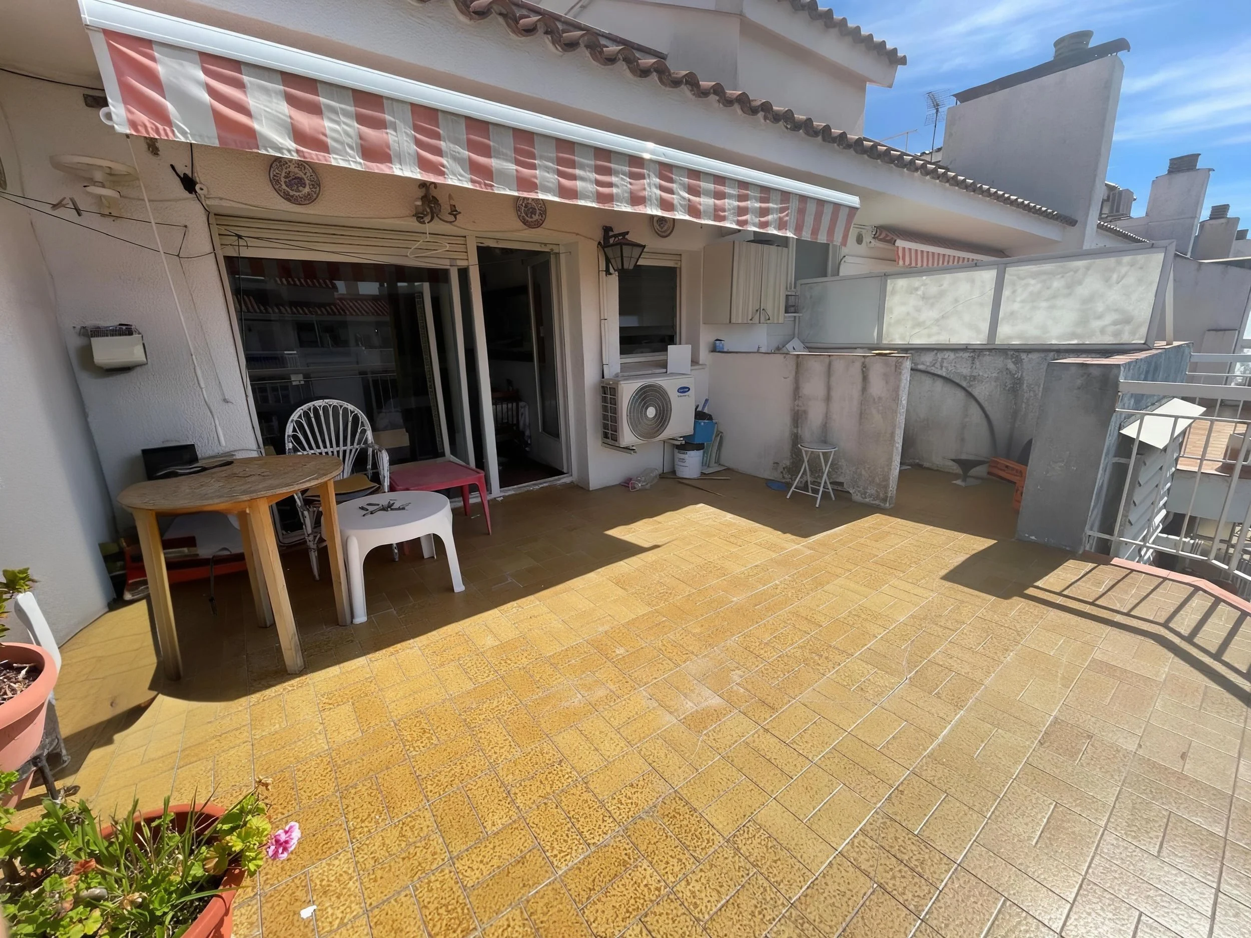 Terraza de un apartamento con piso de azulejos amarillos, algunas plantas en macetas, y muebles como una mesa de madera, sillas blancas, un tambor y una banqueta pequeña, con una vista hacia el cielo y edificios cercanos.