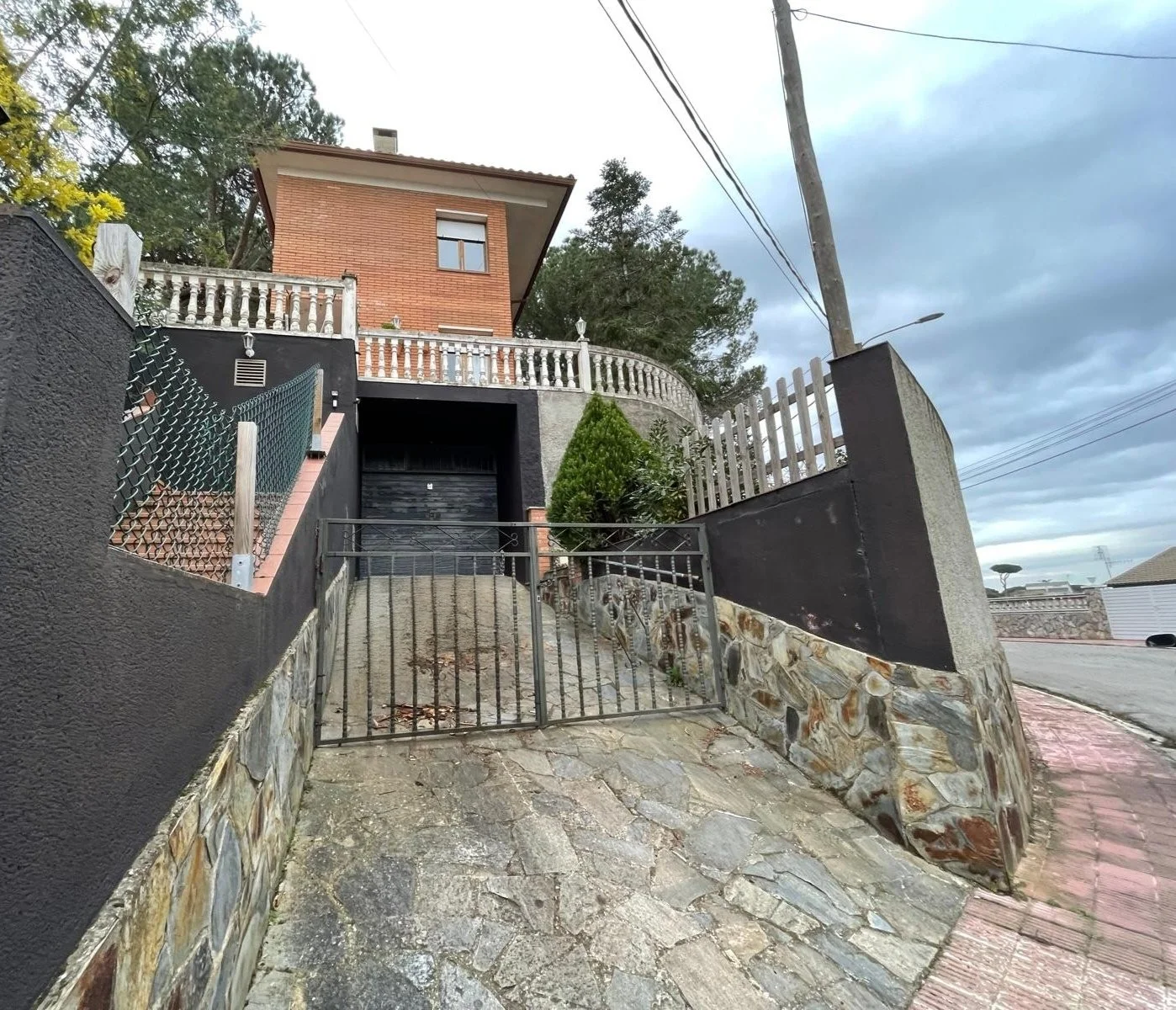 Casa de dos pisos con fachada de ladrillo y muro negro, cerca de una pendiente con rejas metálicas, barandales de piedra y un camino de piedra y concreto.