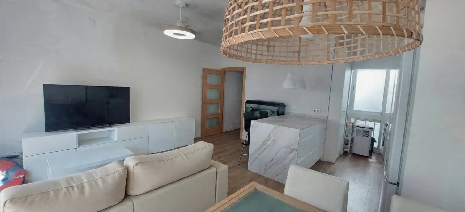 Sala de estar y cocina con mesa de comedor, televisor en mueble blanco, sillón beige, lámpara colgante de mimbre, puerta de madera, ventana con balcón y aire acondicionado.