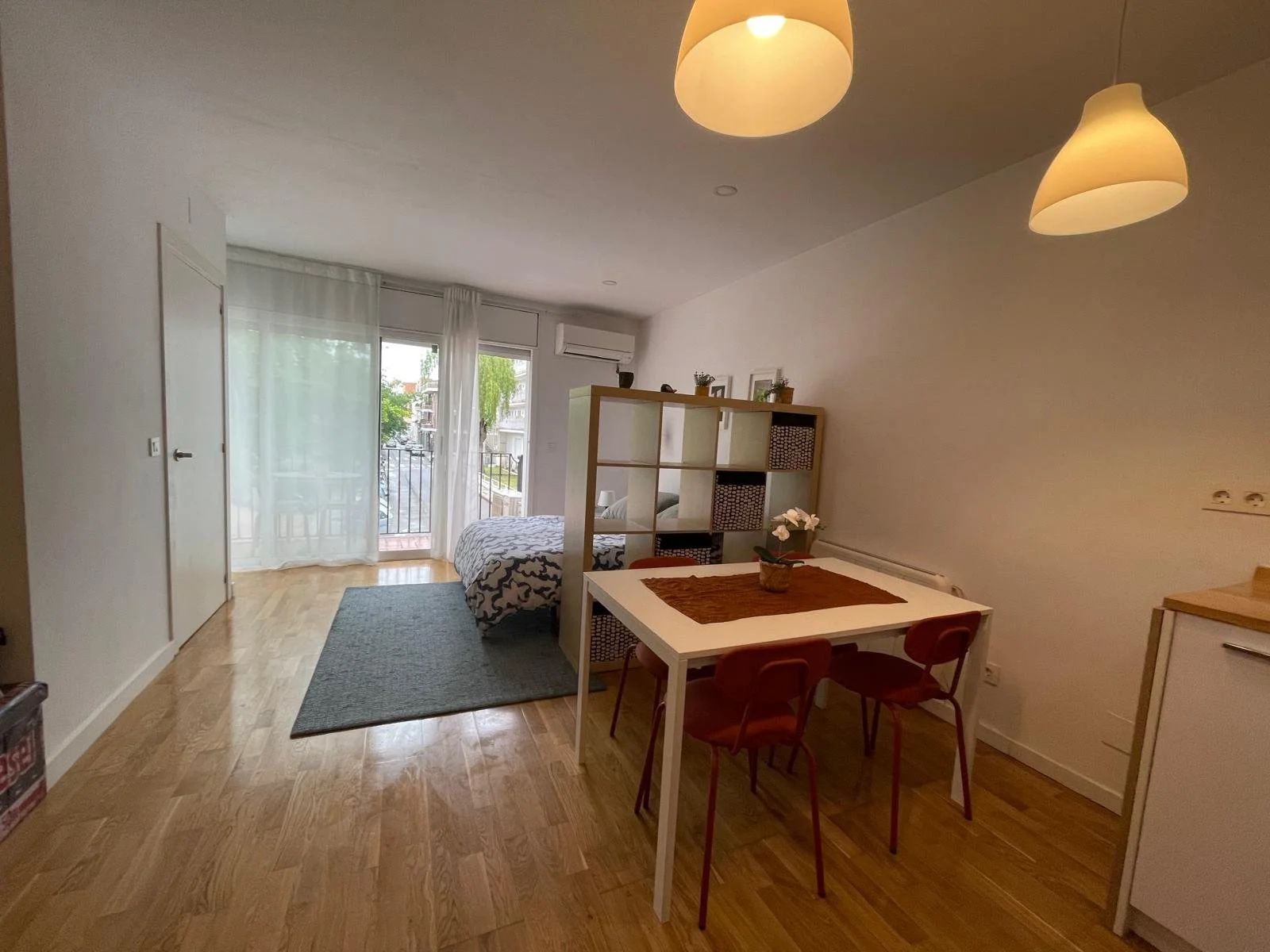 Interior de un apartamento con piso de madera, vista hacia una cama al fondo, una sala con cortinas blancas y balcón, comedor con mesa blanca y sillas rojas, lámparas en el techo, y una separación de estantería