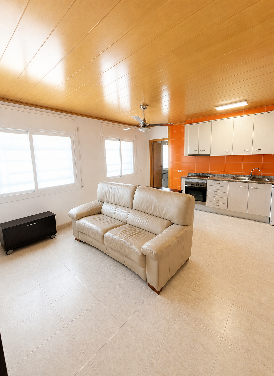Sala de estar con sofá beige, ventana con persianas, muebles de cocina y ladrillos naranja en la pared.