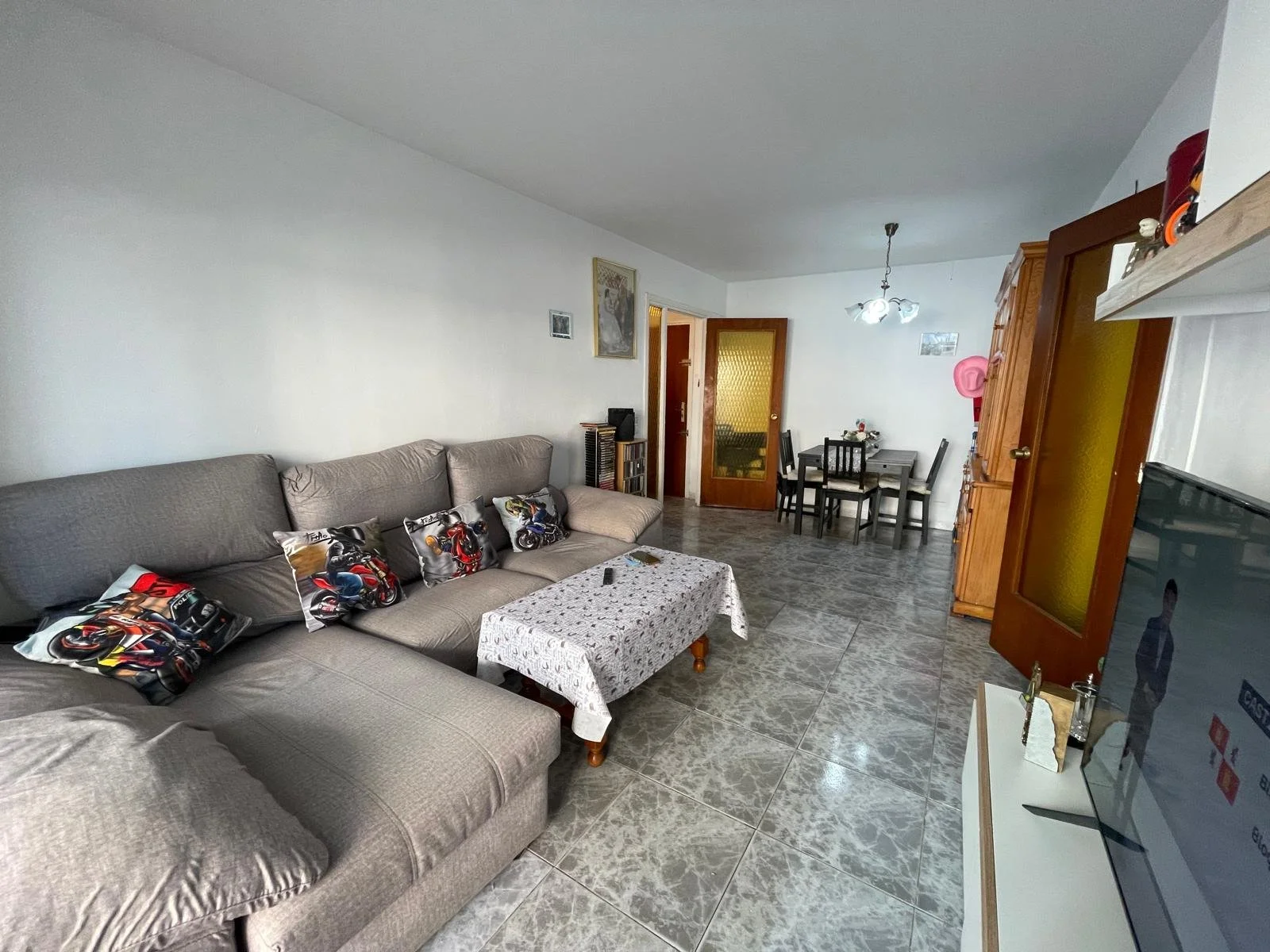 Sala de estar con sofá gris y cojines de motos, mesa de centro con colcha blanca, decoración sencilla, comedor con mesa y sillas, televisor y puerta de madera con cristales, piso de cerámica y paredes blancas.