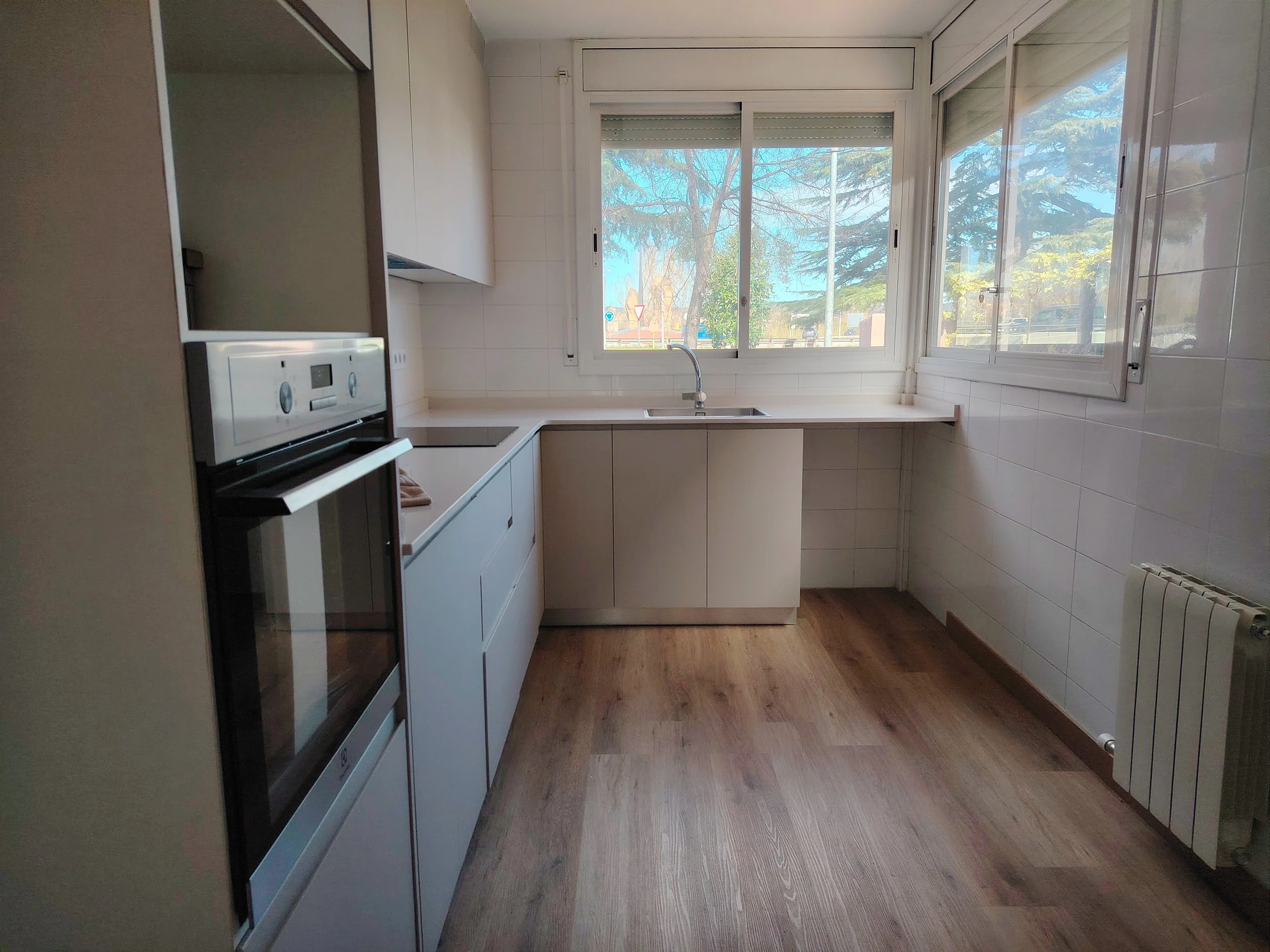 Cocina moderna con ventanas grandes, armarios blancos, horno y suelo de madera
