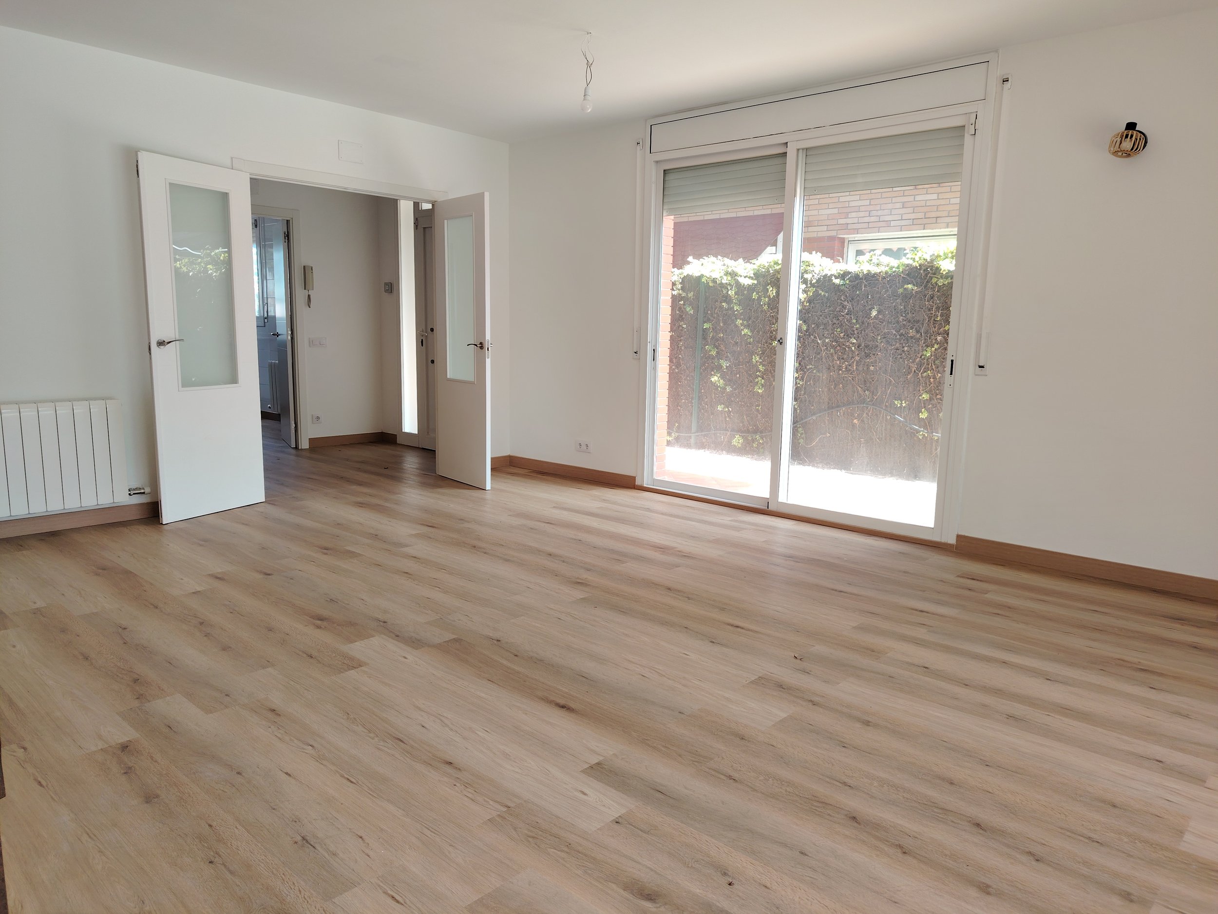 Sala vacía con piso de madera clara, ventanas grandes con vista a un jardín con arbustos, puertas abiertas que conducen a otras habitaciones, paredes blancas, iluminación natural.