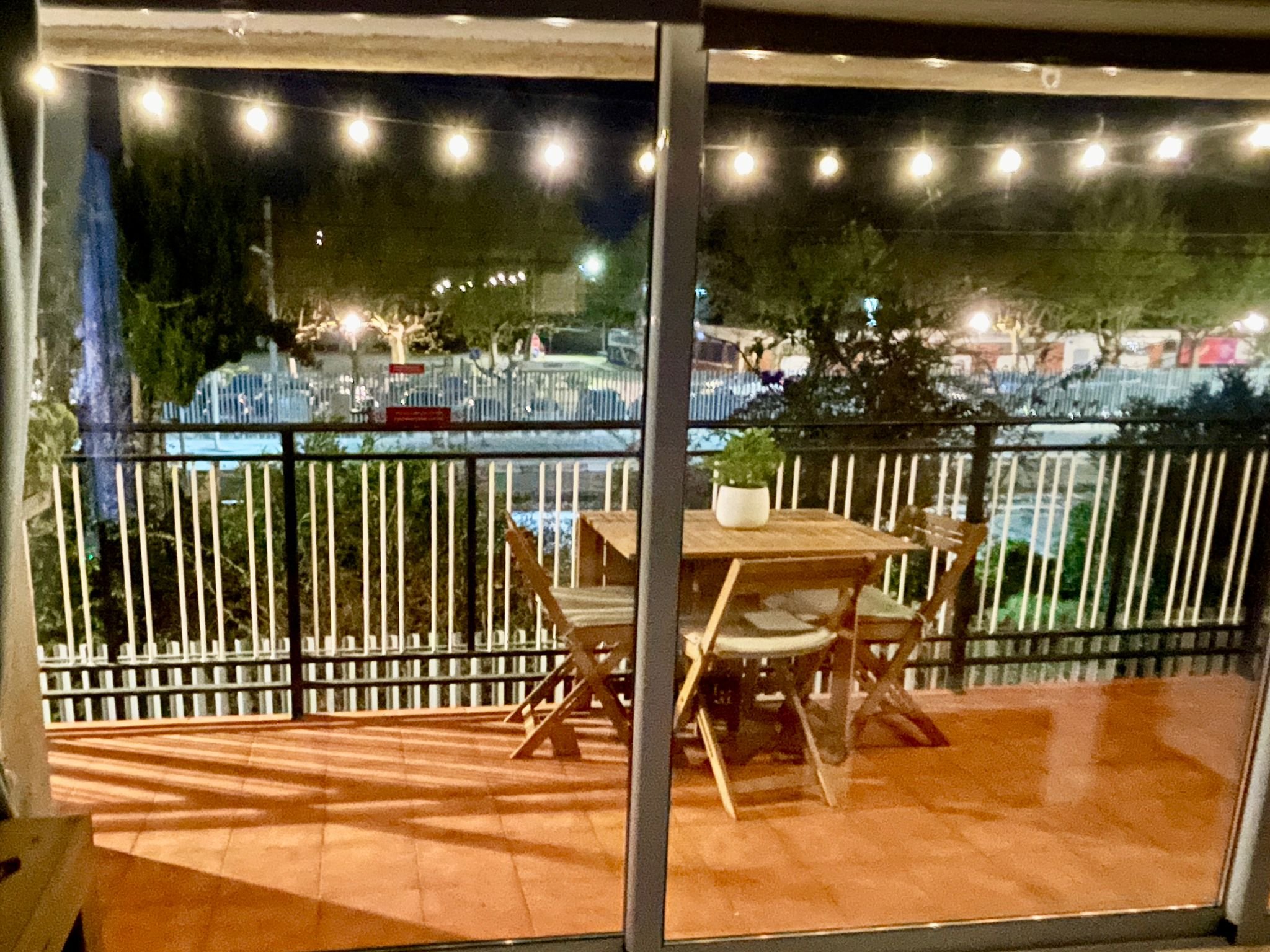 Vista de un balcón con mesa y sillas de madera, con una vista de la calle y árboles bajo luces nocturnas.