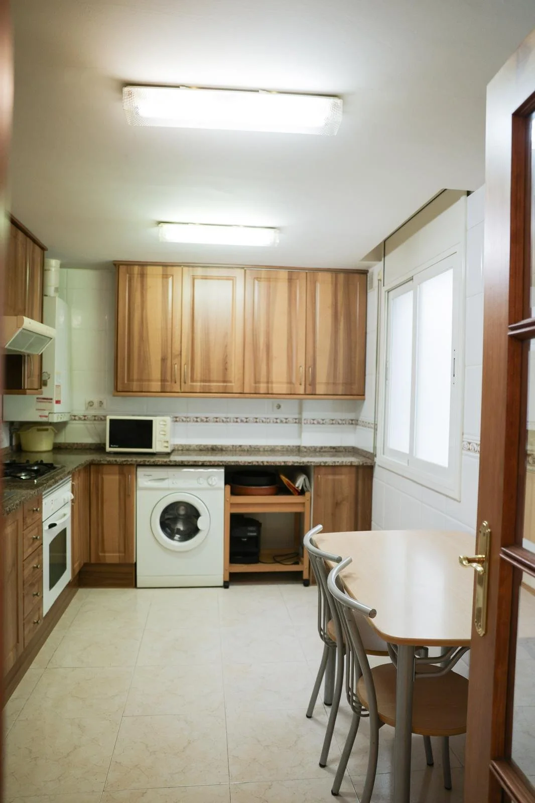 Cocina con gabinetes de madera, microondas, lavadora y mesa con sillas.