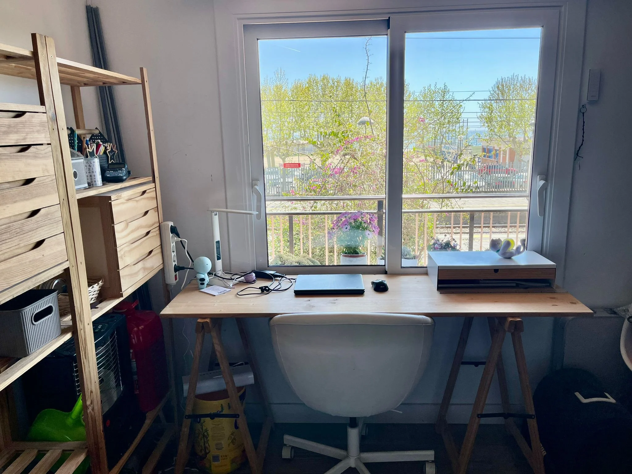 Un escritorio de madera con una laptop, un ratón y una taza. Frente a una ventana grande con vista a árboles y edificios. A la izquierda, estanterías de madera con objetos de oficina y plantas en la ventana.