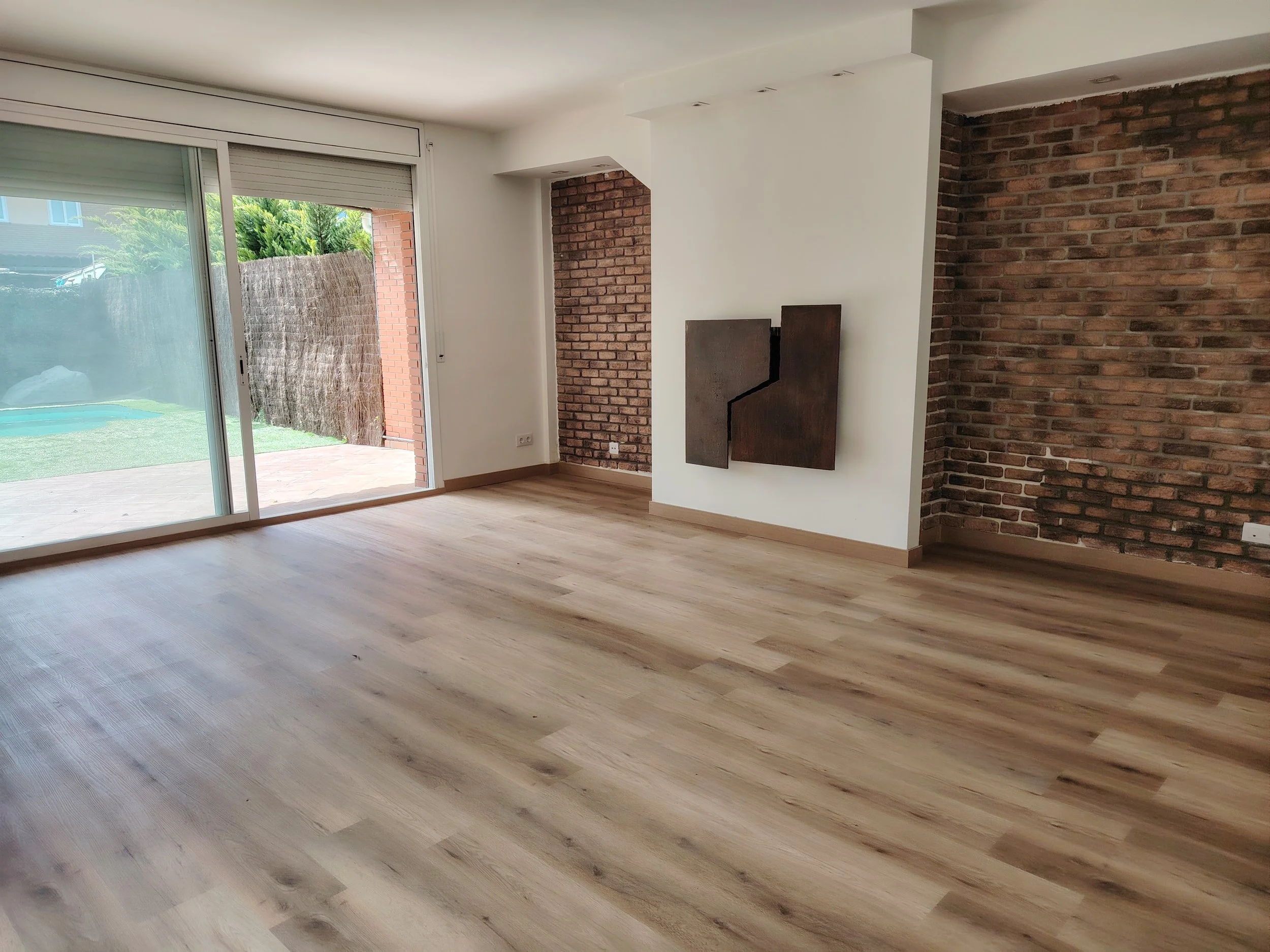 Sala vacía con piso de madera clara, paredes de ladrillo expuesto y blanca, con puerta corrediza de vidrio que da a un jardín con piscina.