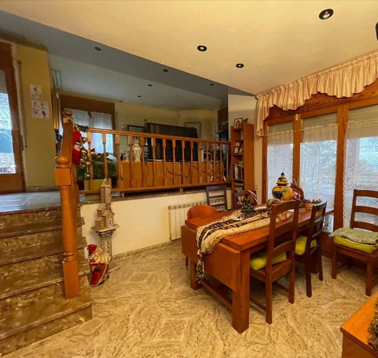 Sala de comedor con mesa de madera, sillas, grandes ventanas con cortinas, decoración navideña, escaleras de mármol, paredes con cuadros y estanterías, ambiente acogedor y cálido.