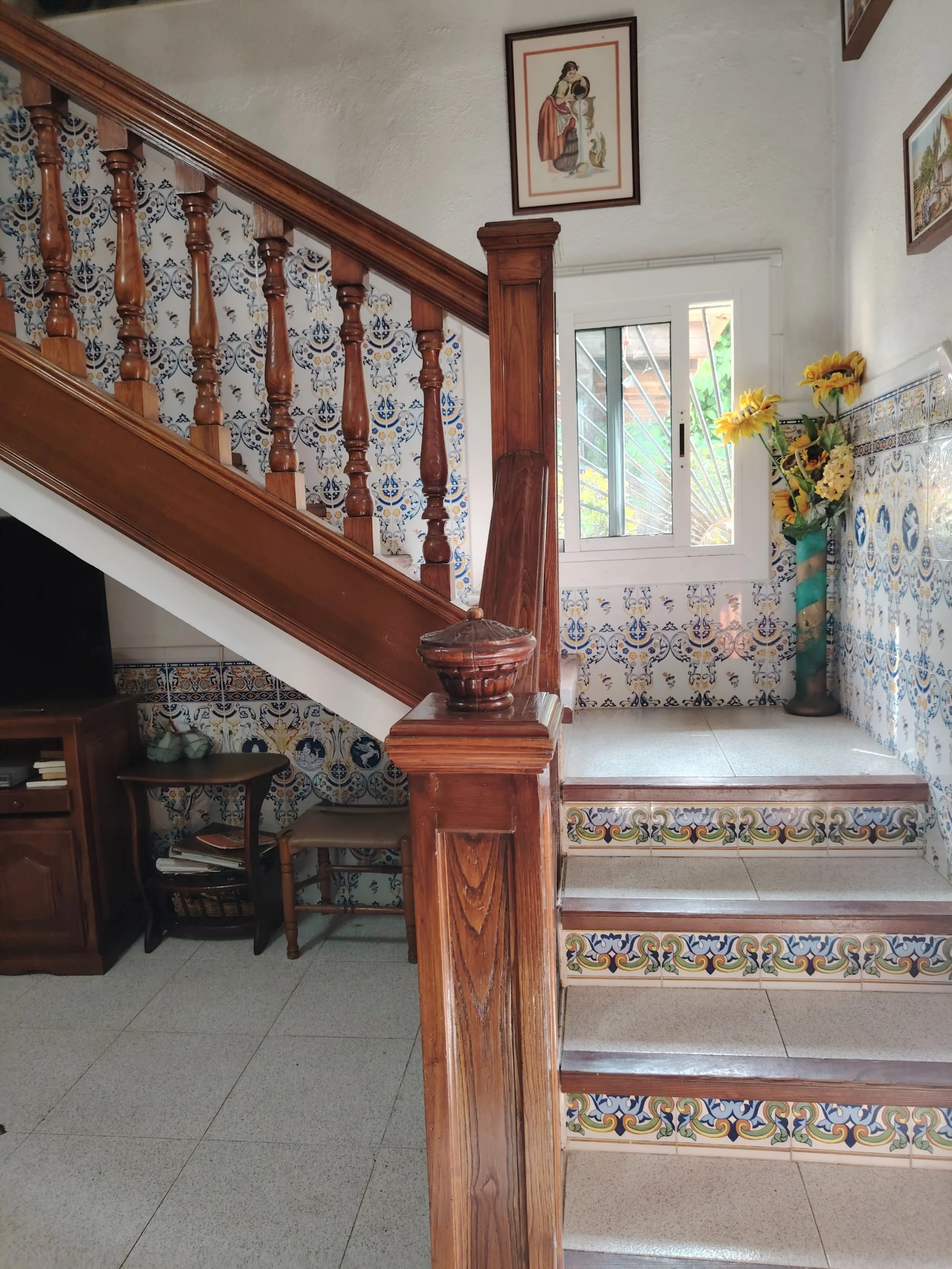 Interior de una casa con escaleras de madera decoradas con azulejos coloridos y flores en una maceta de pared.