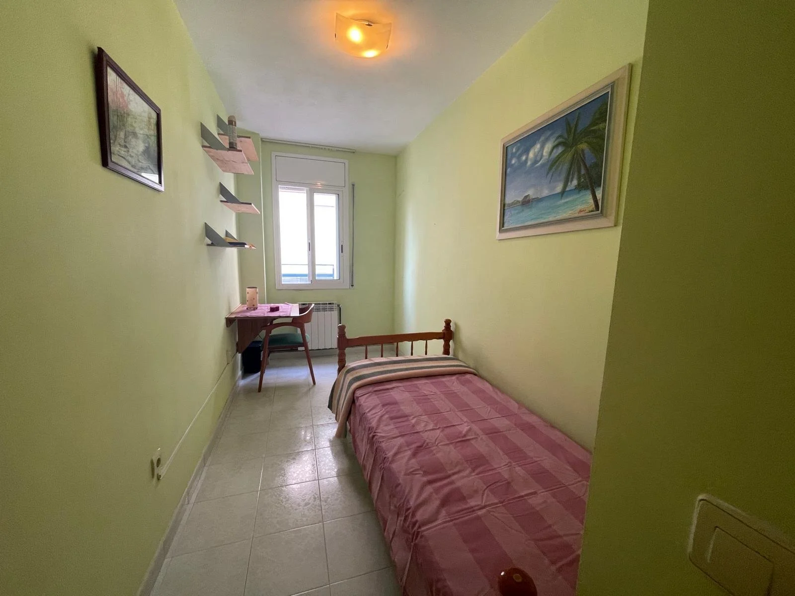 Habitación pequeña con pared verde, cama sencilla con cobertor a rayas, ventana, estanterías, y cuadros con temática marítima.