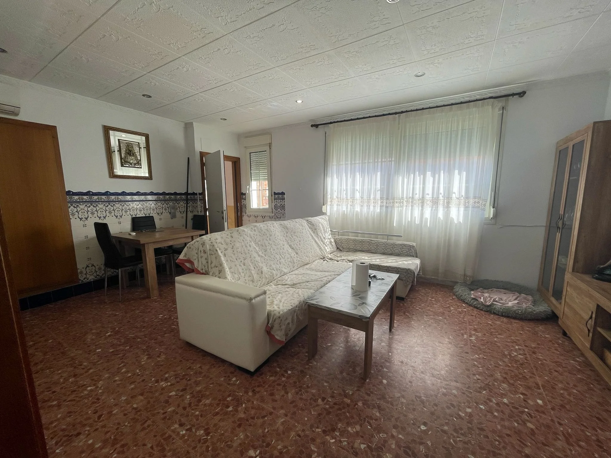 Sala con sofá, mesa, cortinas blancas, pared decorada con azulejos y cuadro en la pared, y piso de terrazo