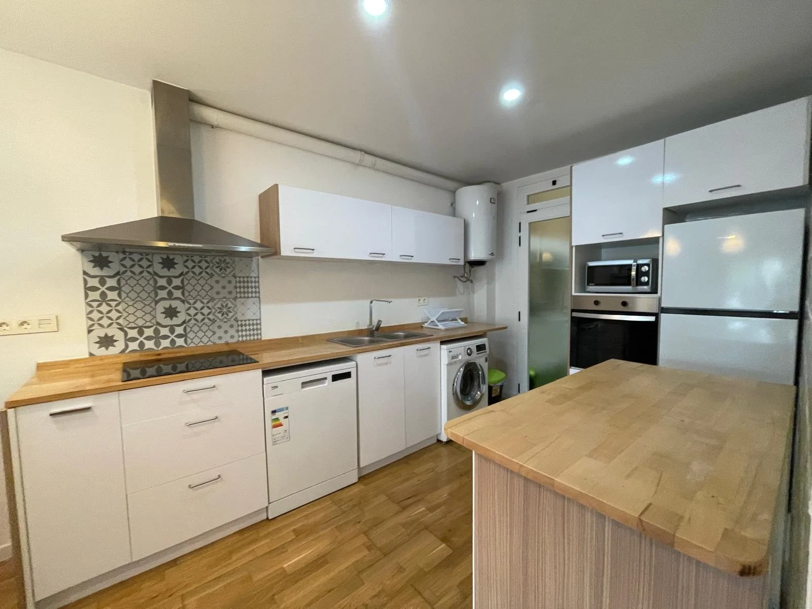 Cocina moderna con gabinetes blancos, estufa eléctrica, campana extractora, fregadero, lavavajillas, lavadora y refrigerador. Encimera de madera y pared decorativa de azulejos con patrones en gris y blanco. Mesa de madera