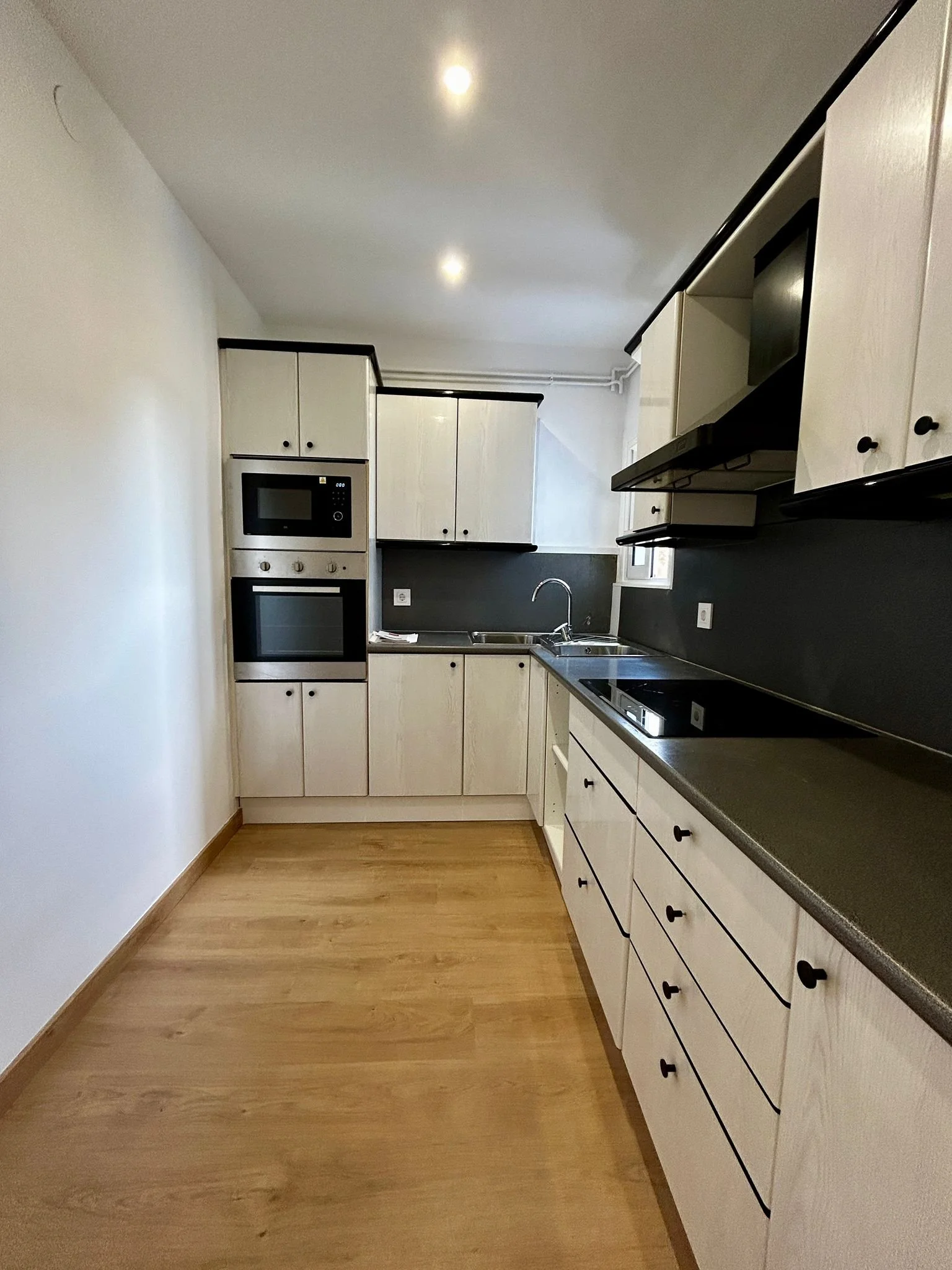 Cocina moderna con muebles blancos, estufa eléctrica, sobres grises, lavabo y paredes blancas, con madera en el piso y paredes negras.