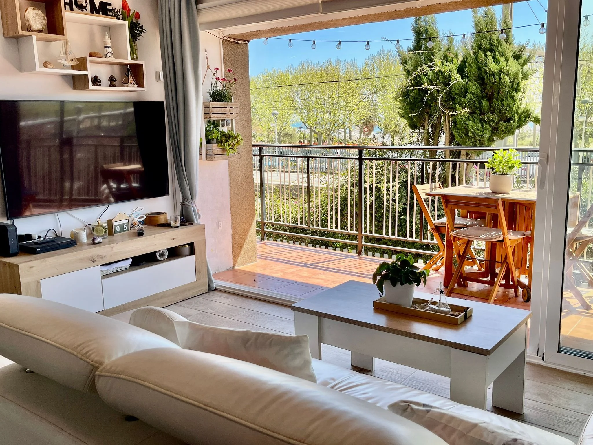 Sala de estar con sofá blanco, mesa de centro, televisor montado en la pared, y balcón con mesas de madera, plantas y árboles verdes en un día soleado.