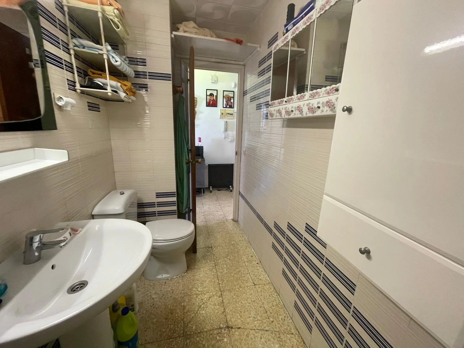 Un baño pequeño con lavabo, inodoro, estantes con ropa y artículos, y un armario grande, con puerta abierta que revela una habitación con cuadros en la pared al fondo.