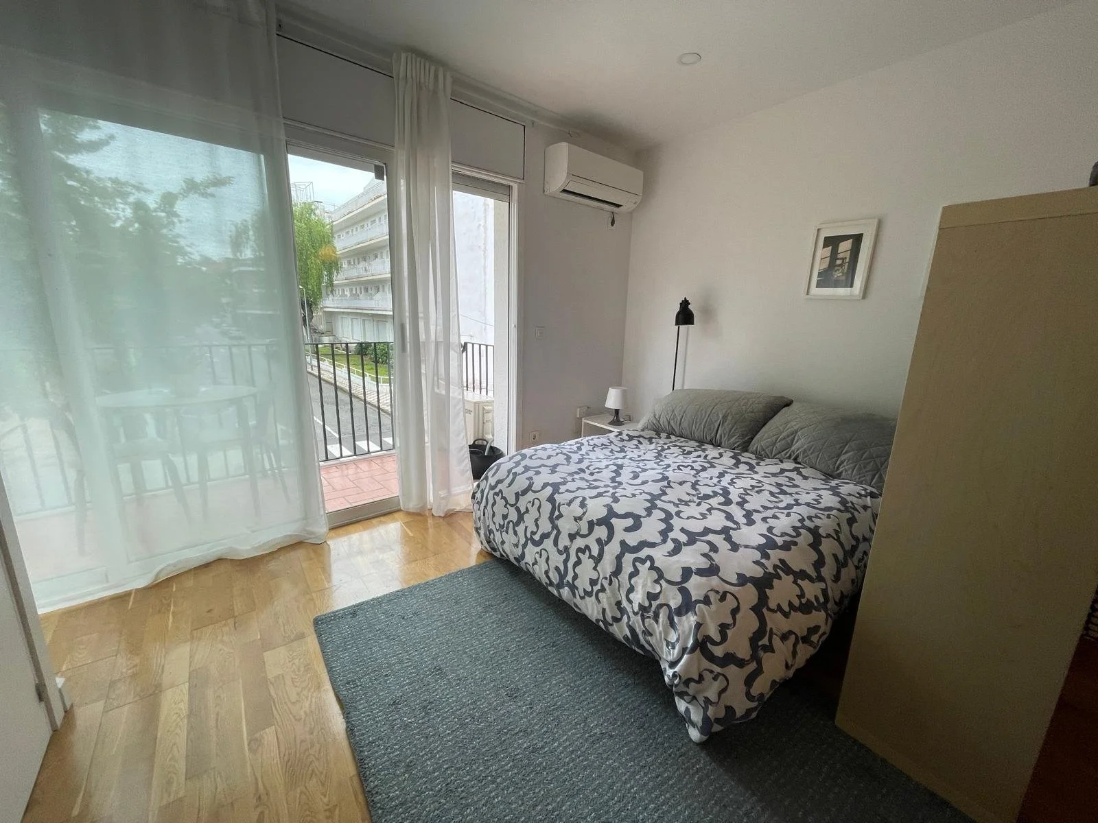 Habitación con cama doble, cortinas blancas, vista a un balcón con barandilla, aire acondicionado en la pared, lámpara de lectura en la mesita y un cuadro en la pared.