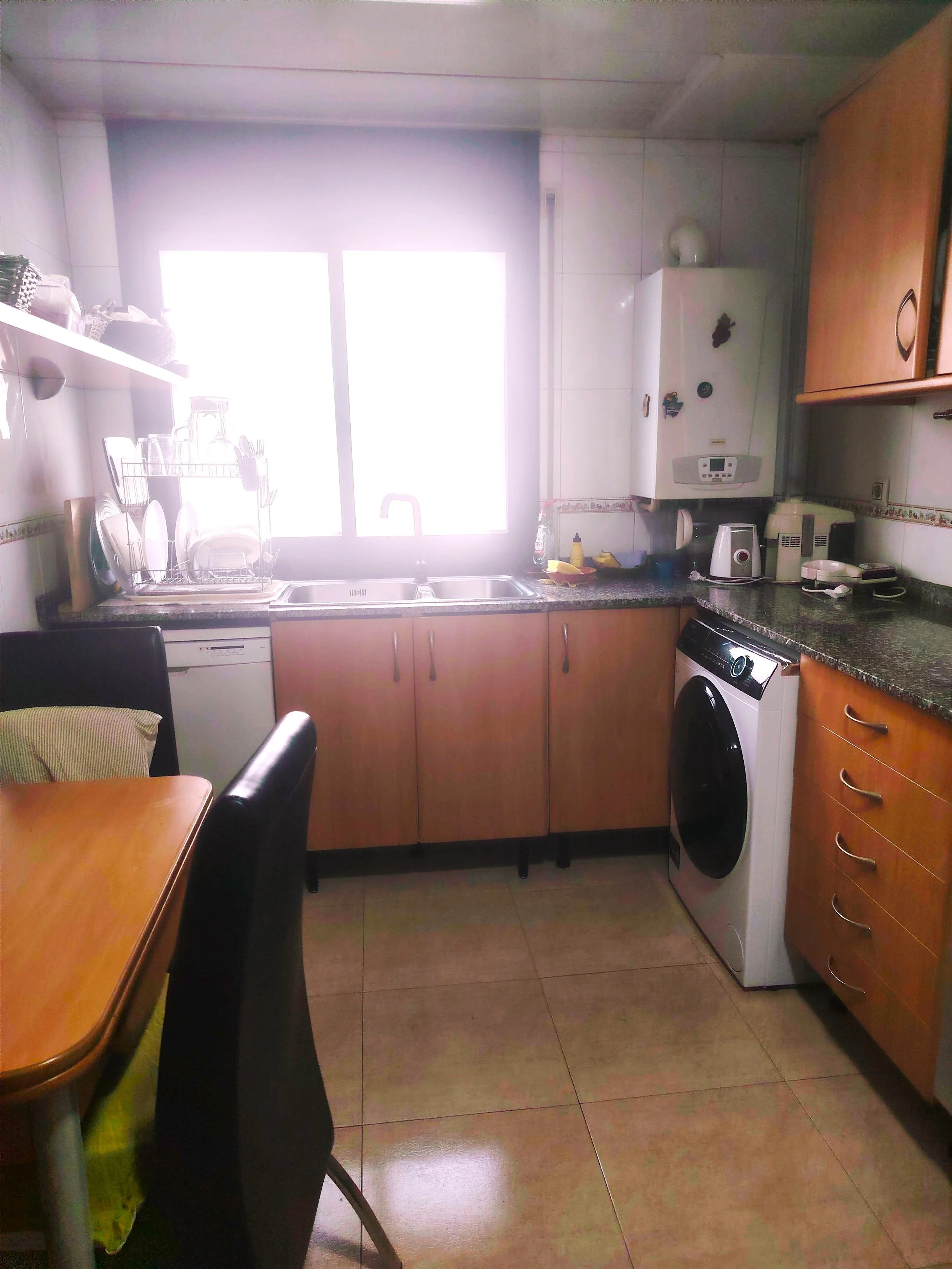 Cocina con fregadero, estante con utensilios y electrodomésticos como cafetera y lavadora. Ventana grande que deja entrar luz.