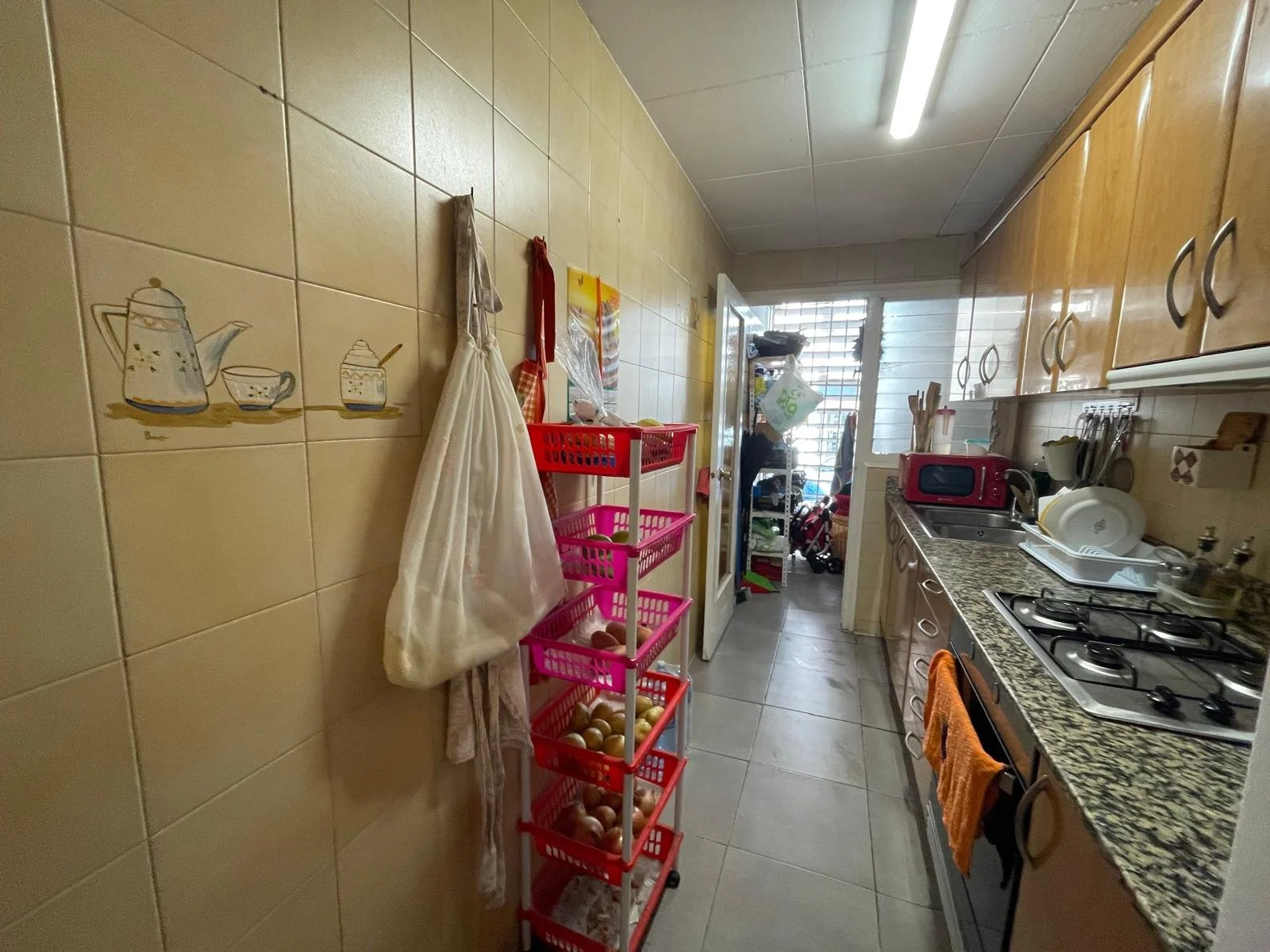 Cocina con gabinetes de madera, estufa de gas, encimera de granito, utensilios de cocina, y armario con puertas. En la pared, azulejos decorativos con dibujos de teteras y tazas. En el centro, un carrito con huevos, y al fondo, una puerta con ventana de barrotes y una habitación con objetos y bolsas.