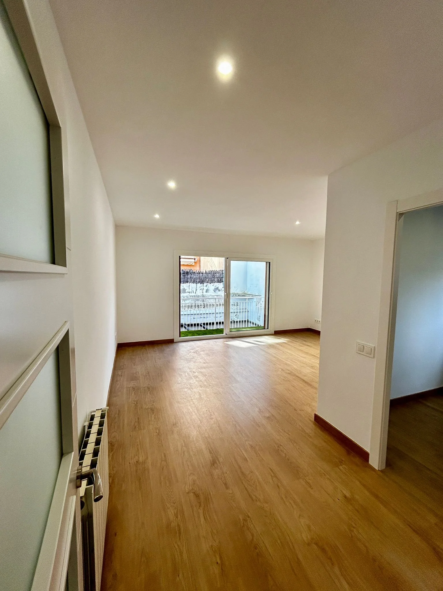 Sala vacía con suelo de madera, paredes blancas, ventanas de cristal y salida a un balcón con barandilla.