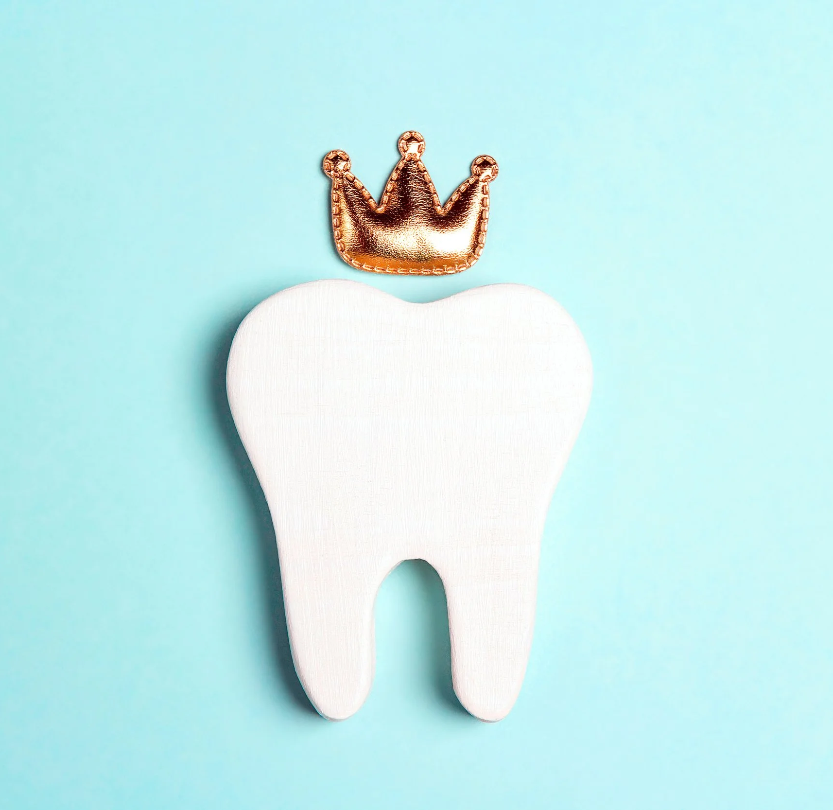 A white wooden tooth cutout with a small gold crown ornament placed on top, set against a light blue background.
