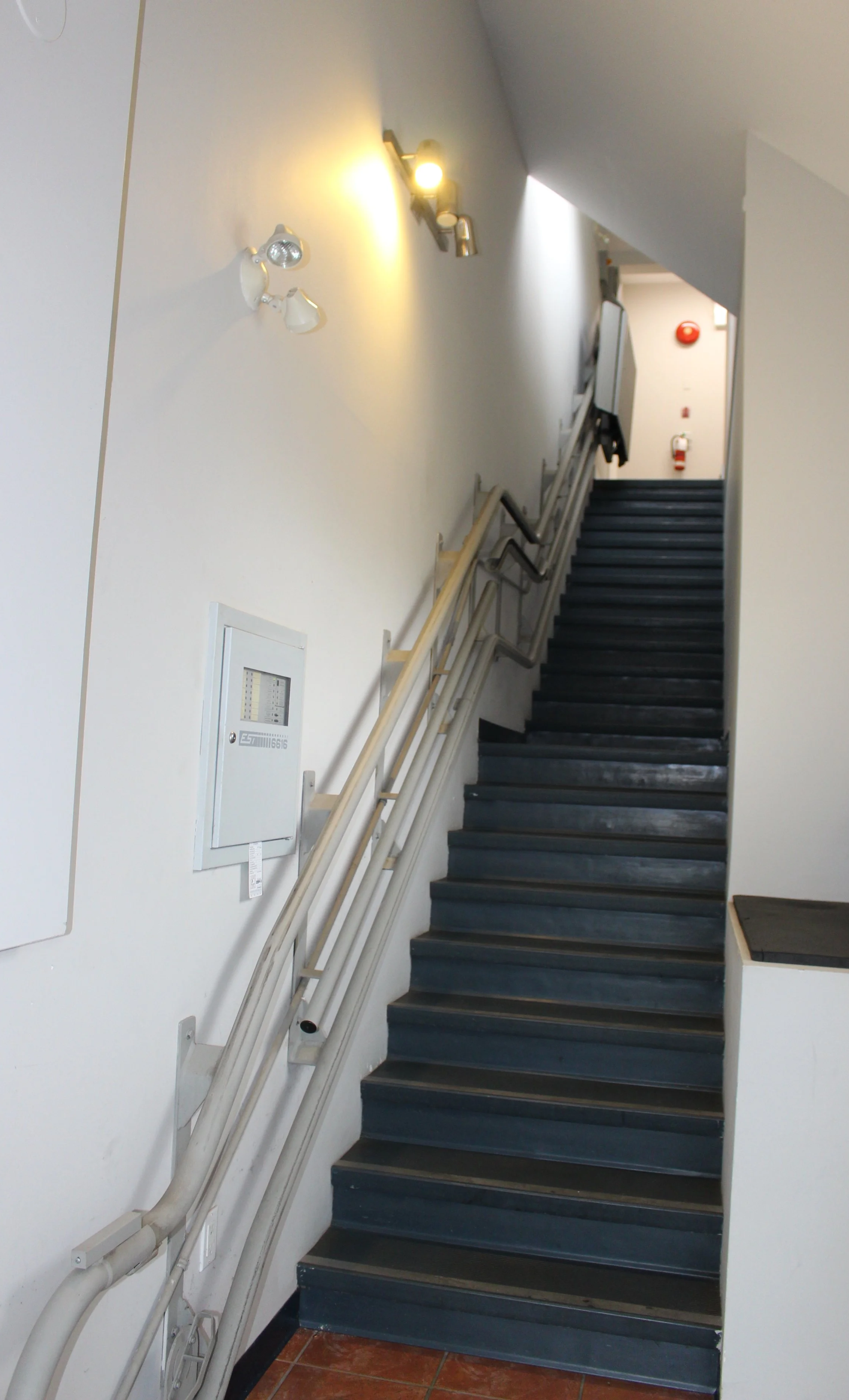 Image of a stairway with a blue railing and dark steps, leading to an upper floor. There are wall-mounted lights and fire safety equipment at the top. The wall is white, and the floor is tiled.