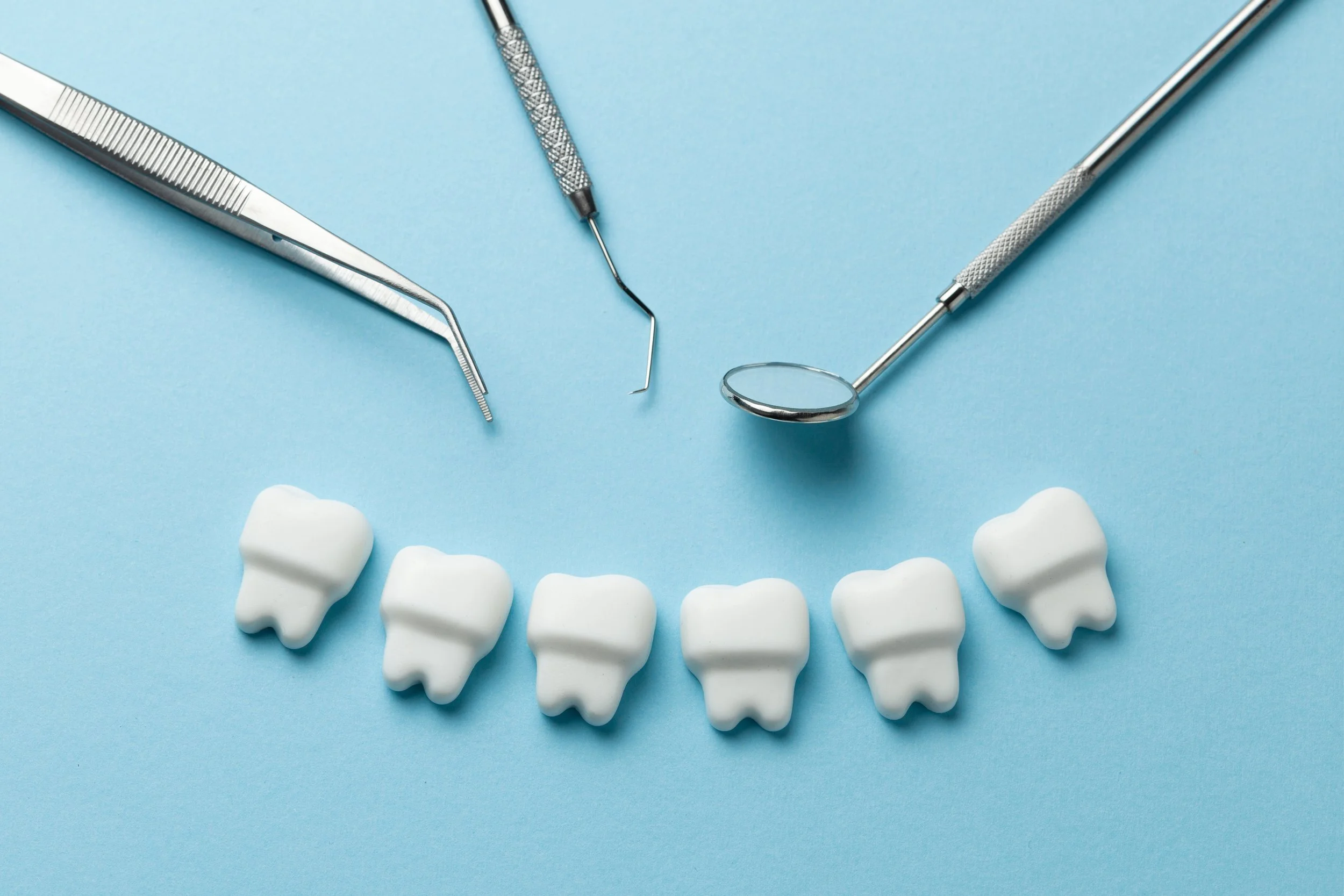Six artificial teeth arranged in a smile, dental tools including a mirror, explorer, and tweezers on a light blue background.