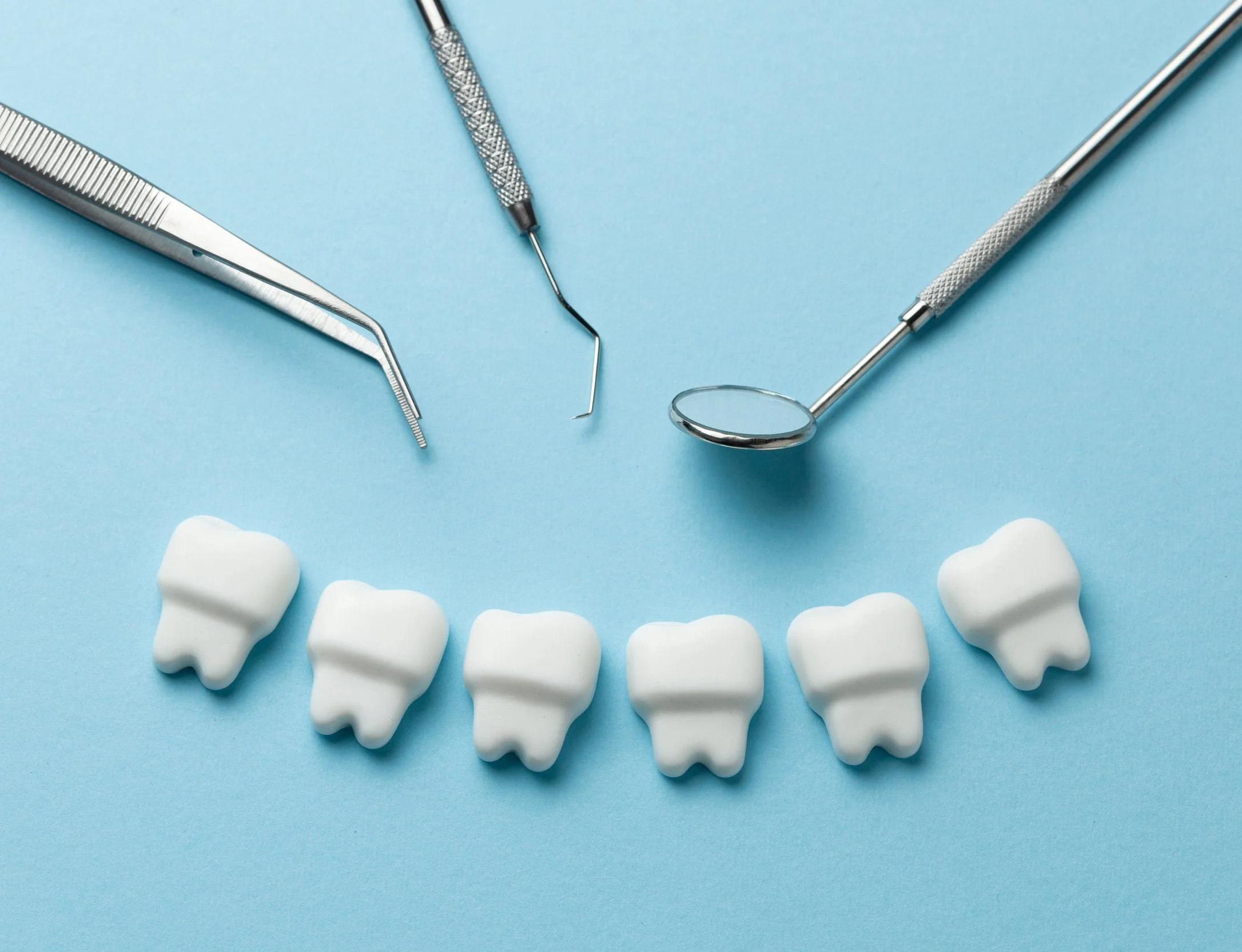 Six white dental prosthetic teeth arranged in a row on a light blue background. Three dental tools, including a mirror, a probe, and a periodontal scaler, are positioned above the teeth.