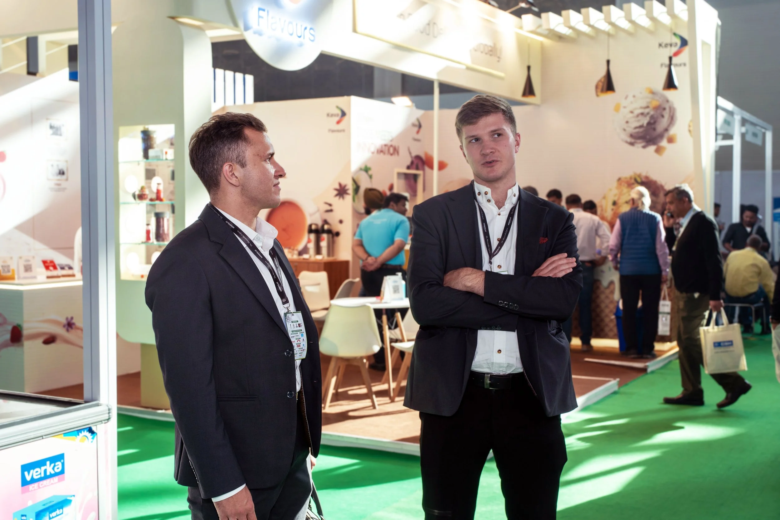 Two young men in business suits chatting at an exhibition booth with a green carpet, other attendees in the background, and food and beverage displays.