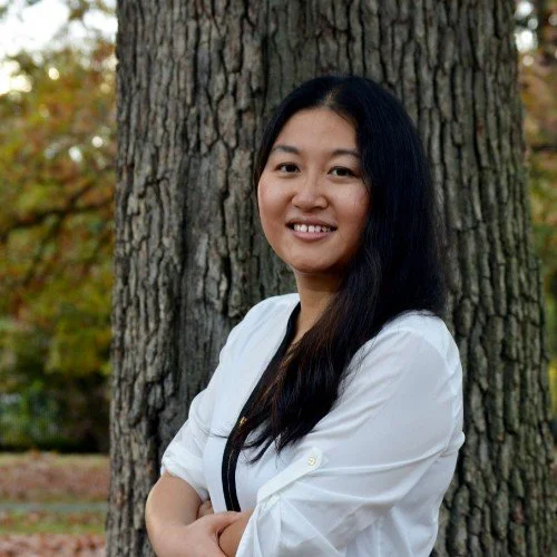 A woman with long black hair smiling and standing outdoors in front of a large tree, with autumn leaves in the background.