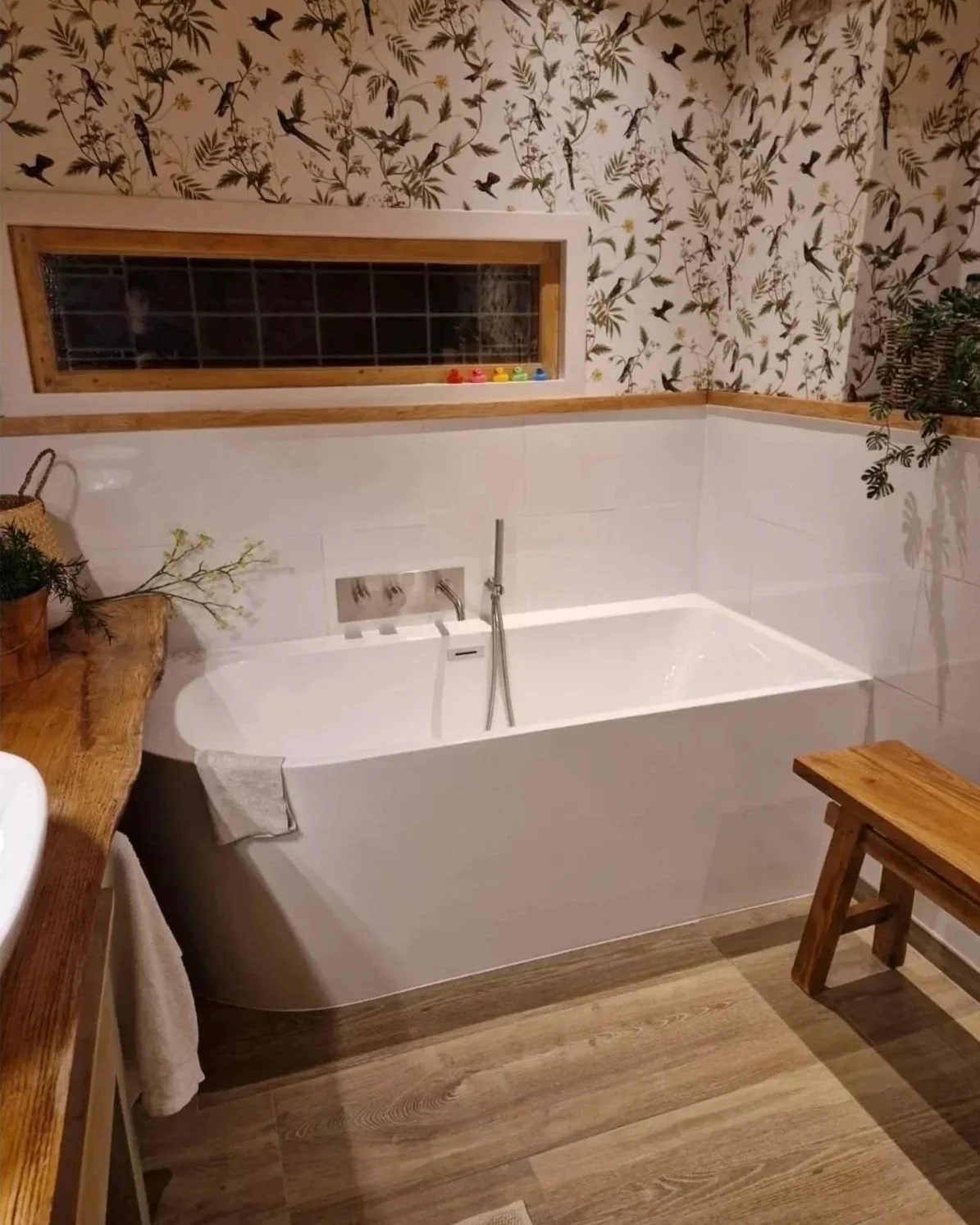 Een badkamer met een ligbad, een houten bank en planten. De muur heeft een decoratief behang met vogel- en bladmotieven.