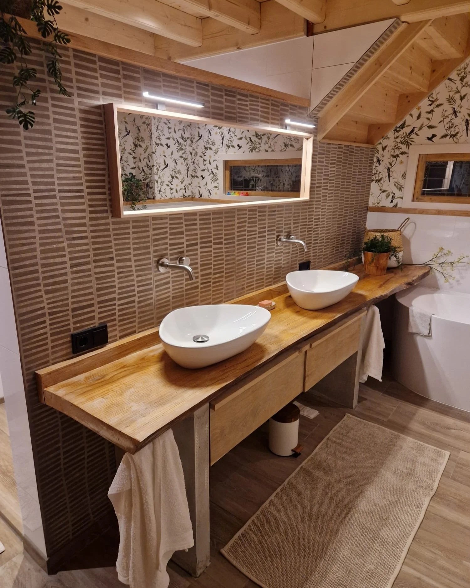 Een badkamer met een houten wastafel met twee waskommen, een spiegel met ingebouwde verlichting, een hennepplant, een badkuip, en een vloerkleed, met een muur van keramische tegels en behang met vogel- en bladmotieven.